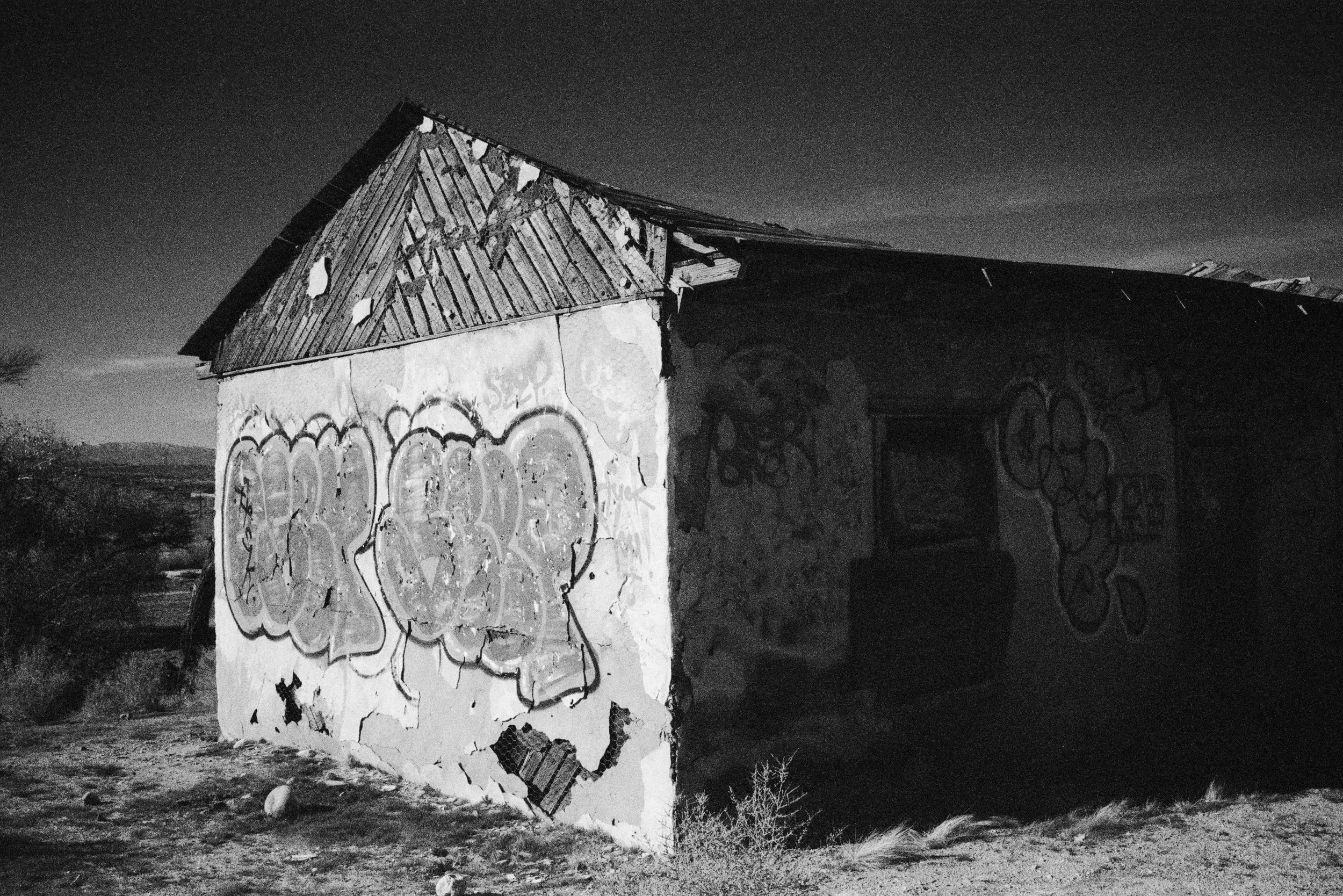 An old, abandoned building with graffiti on the walls, a damaged roof, and a dark interior. It is in a desolate area during dusk or night.