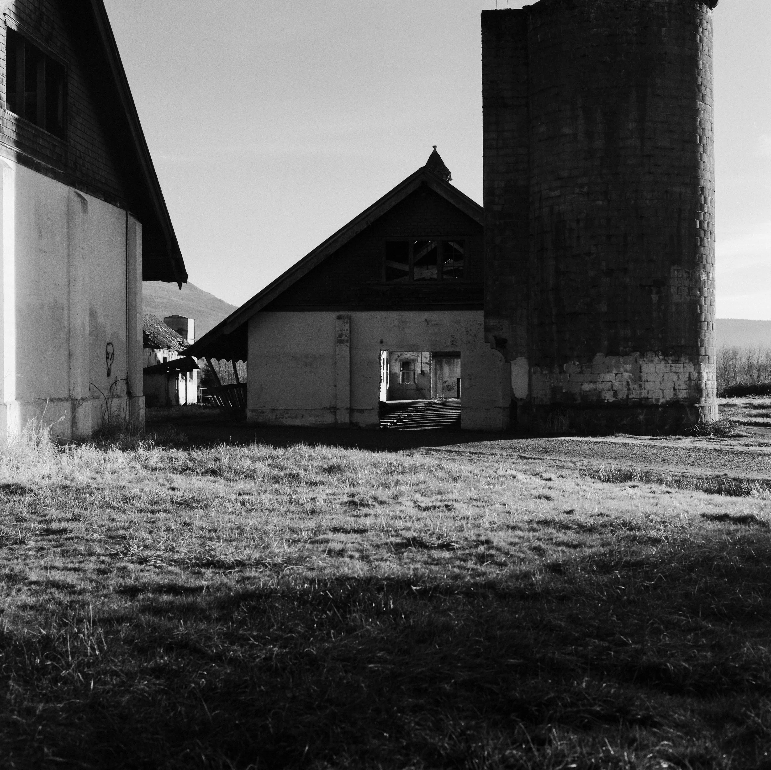 Black and white photo of old, abandoned farm buildings with a silo on the right and a barn with a sloped roof in the center, with grass in the foreground and mountains in the background.
