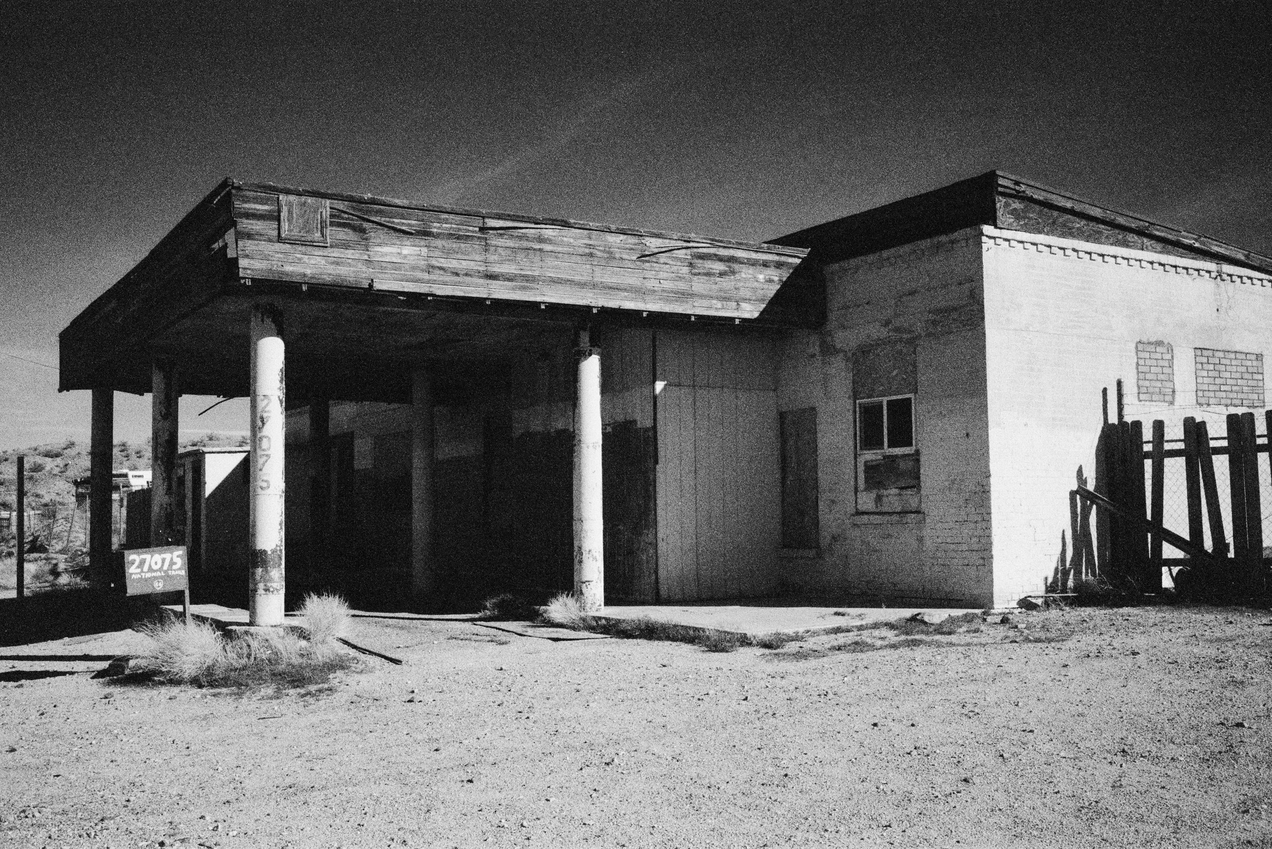 Black and white photo of an abandoned building in a desert landscape. The building has a partially collapsed roof, weathered wooden and brick walls, and overgrown dirt ground with sparse desert vegetation.
