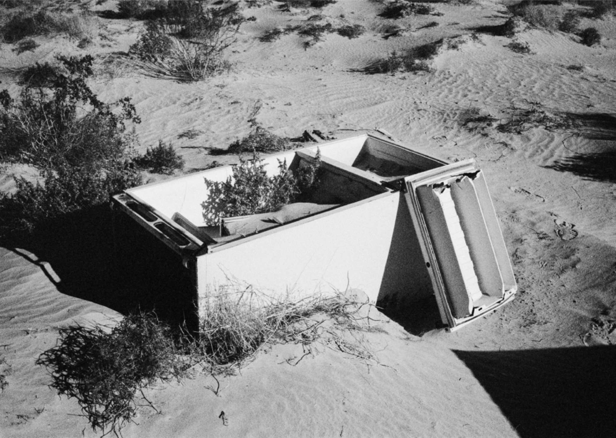 An overturned boat in sandy desert terrain with sparse vegetation and shadows cast on the sand.