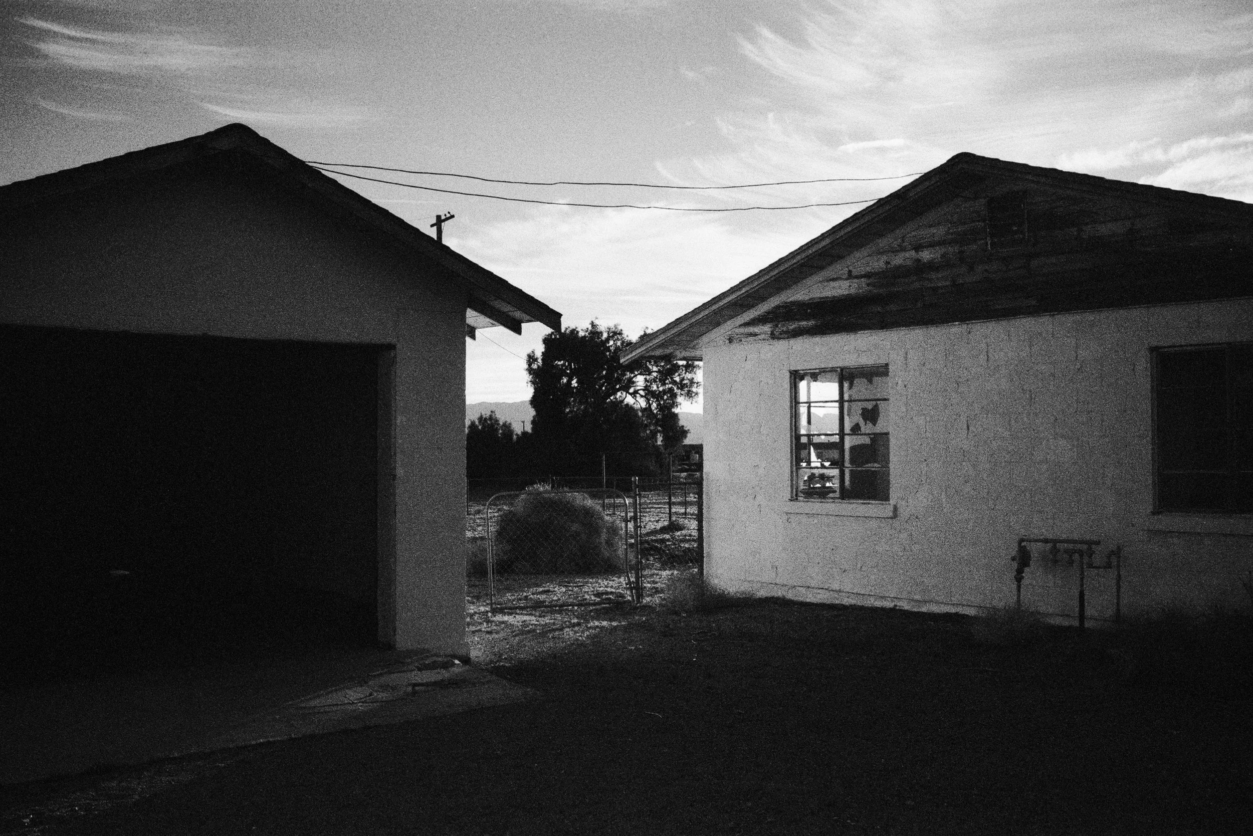 Black and white photo of two houses with a wire fence between them, a tree in the background, and overhead power lines.