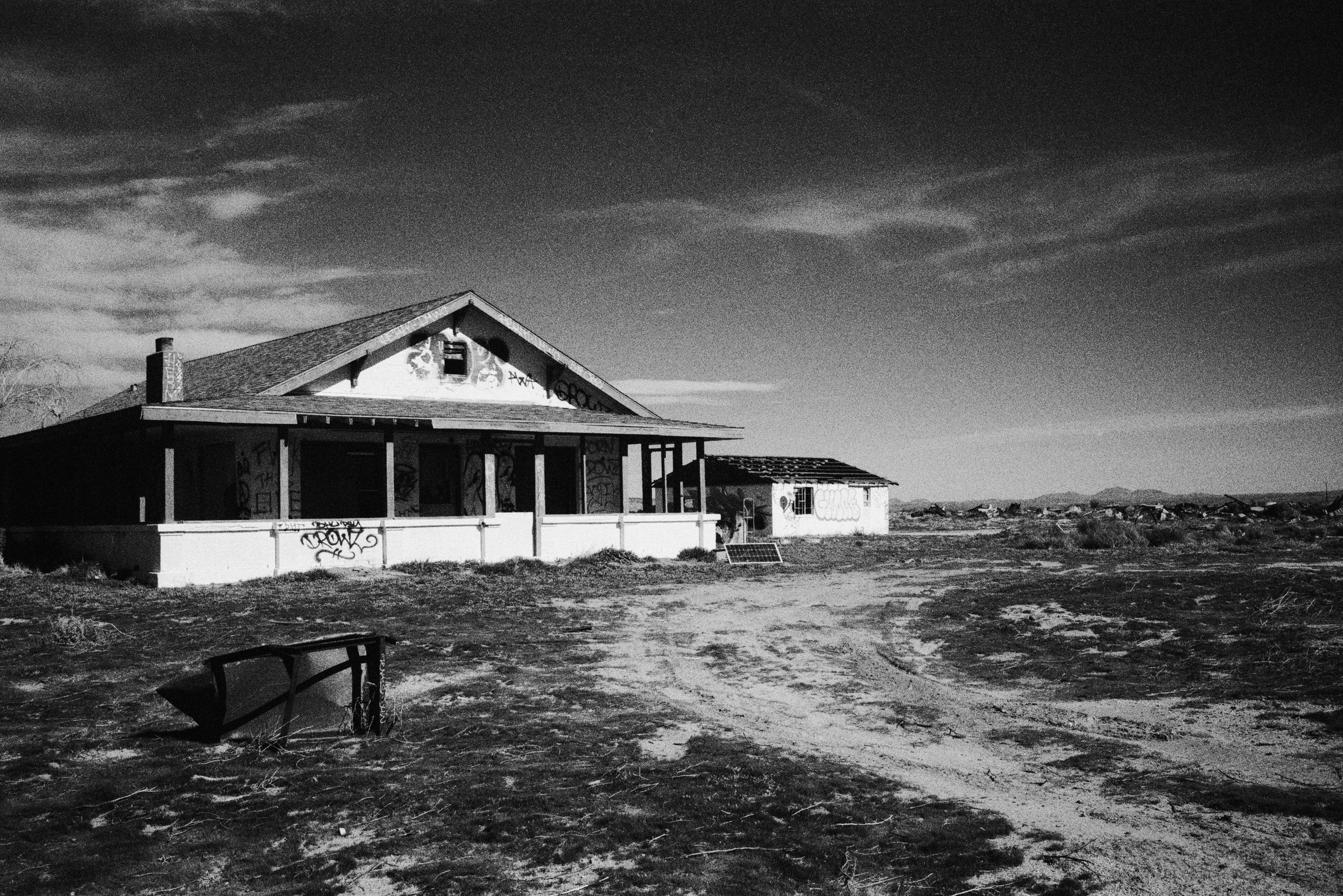 Abandoned house in a desolate, desert landscape with graffiti on the walls and a broken grill in the foreground, under a cloudy sky.