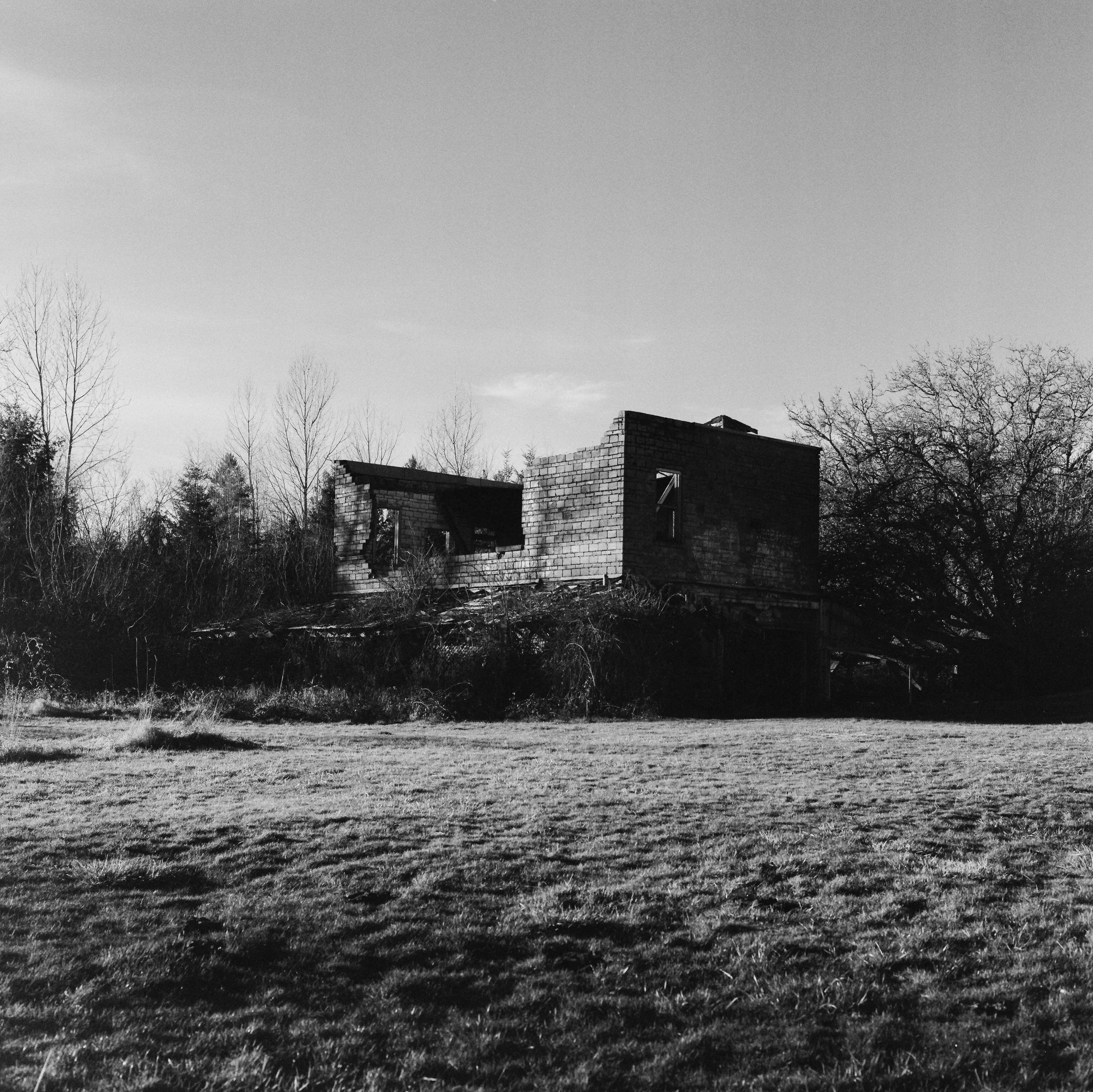 Black and white photo of an abandoned, partially collapsed house on a grassy field with leafless trees in the background.