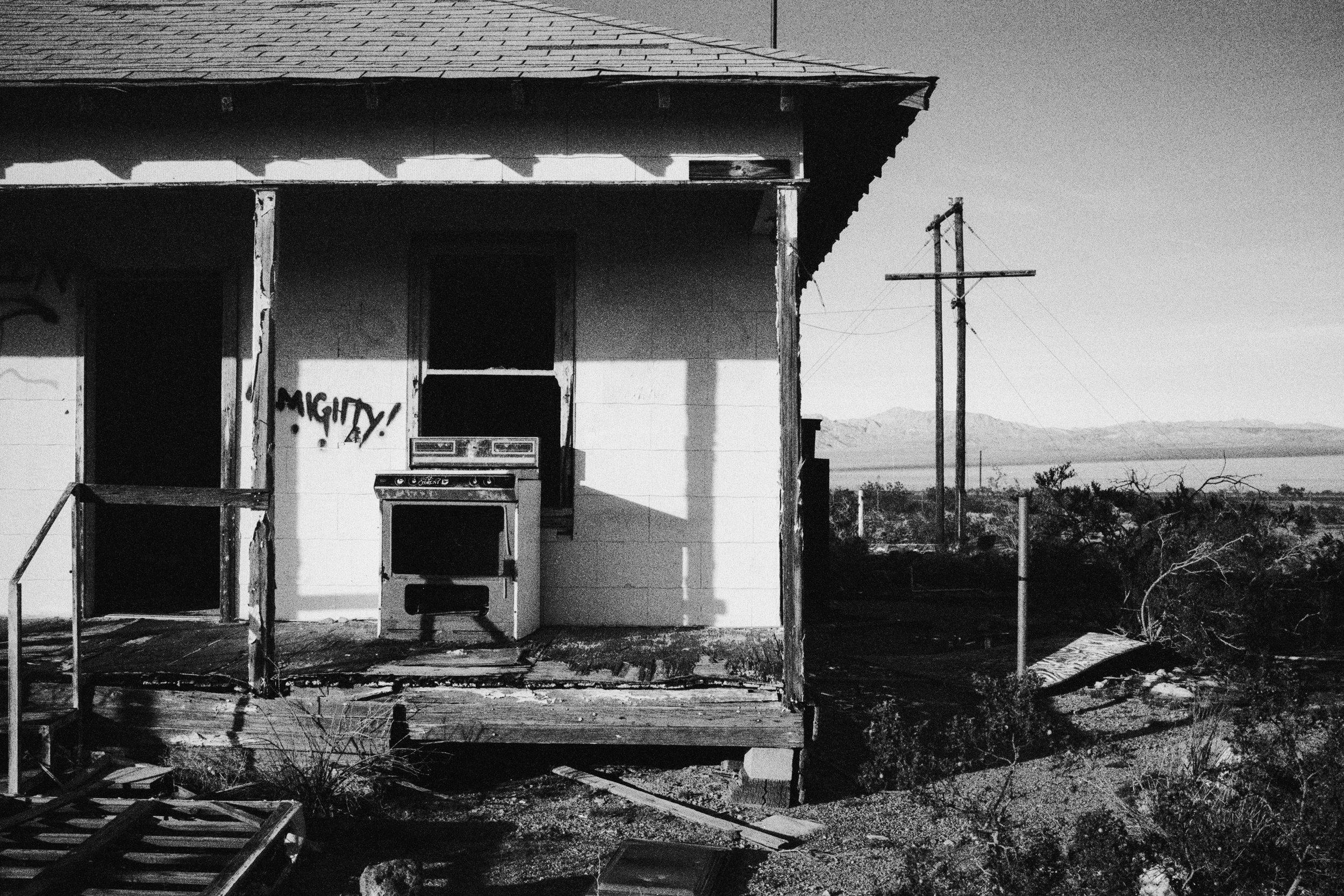 Black and white photo of abandoned house with broken porch, old stove, and graffiti. Desert landscape and power lines in the background.
