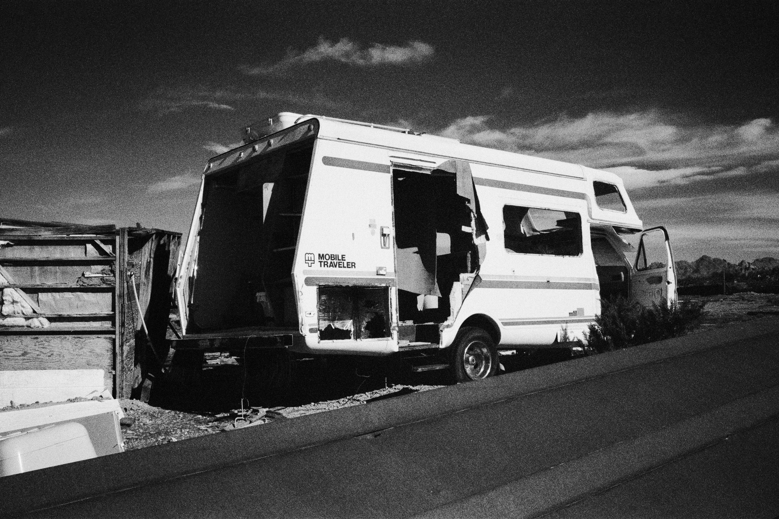 A black and white photo of an abandoned RV trailer parked on the side of a road with a mountainous landscape in the distance.