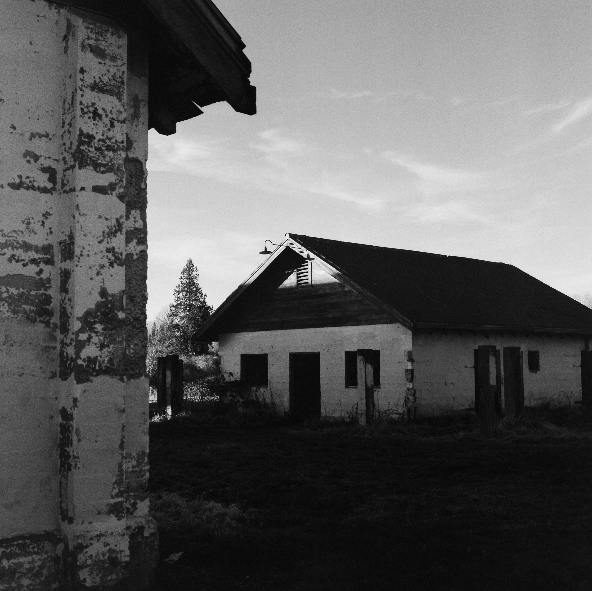 Black and white photo of two old rustic buildings with a tree in the background and a partly cloudy sky.