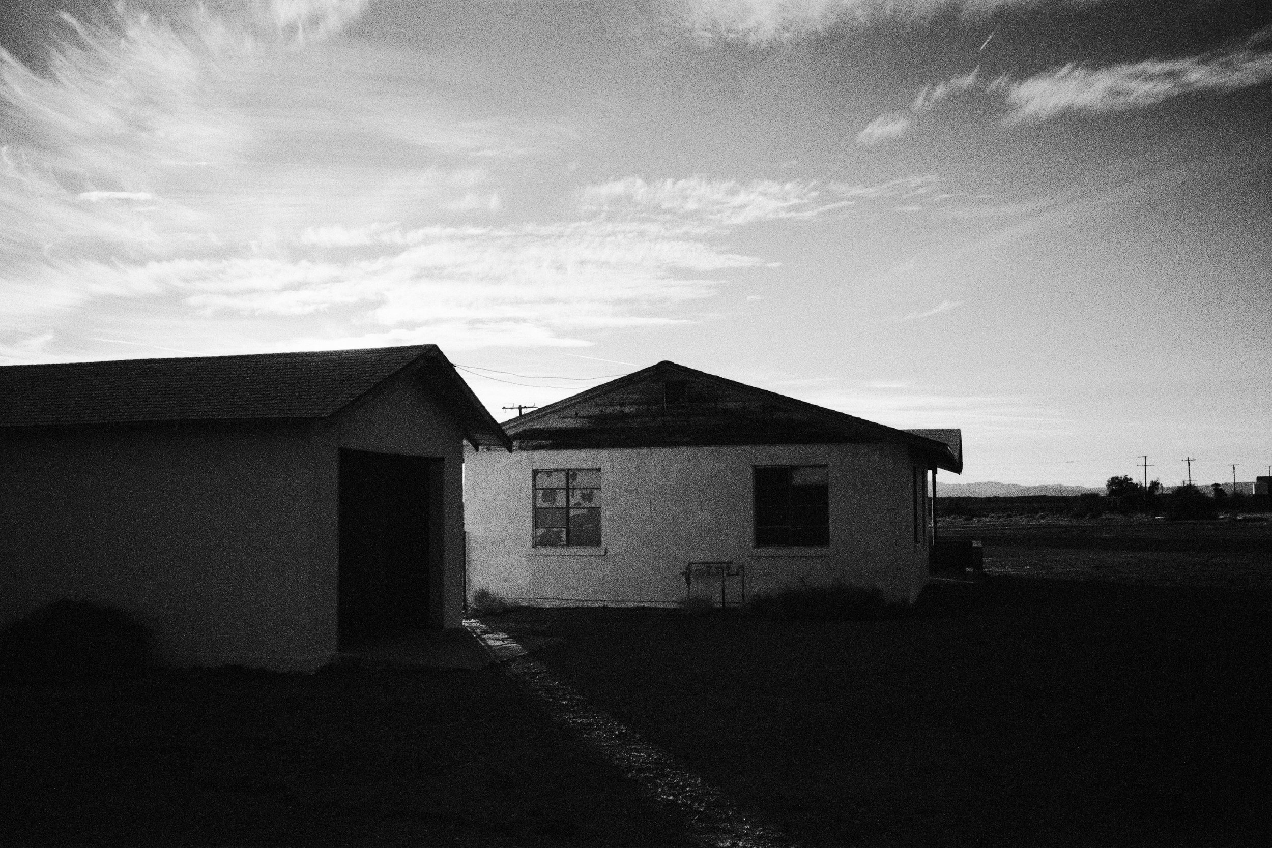 A black and white photo of two houses, one with a gable roof and a garage, under a sky with scattered clouds.