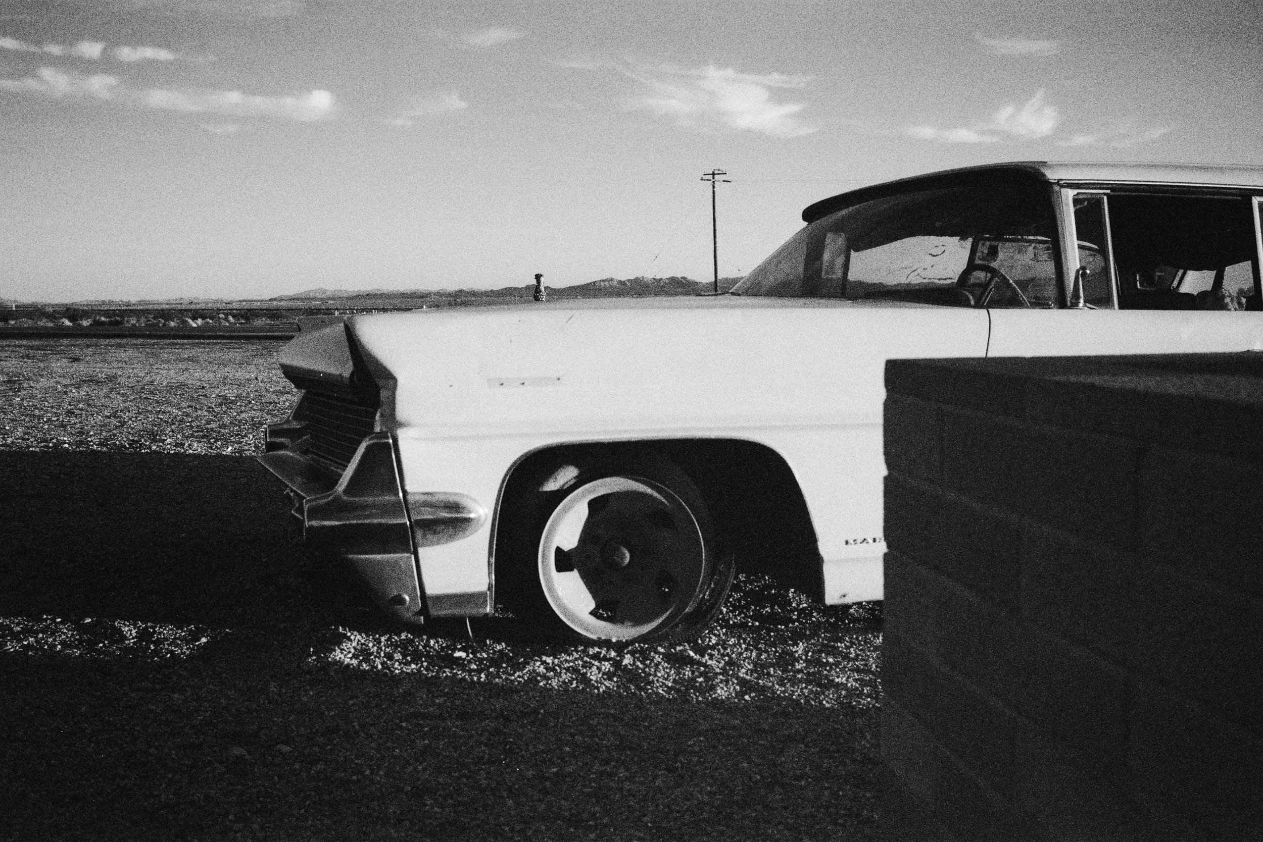 A black and white photo of an abandoned vintage car with missing wheels, parked on a gravel area near an open landscape with a person fishing in the distance.