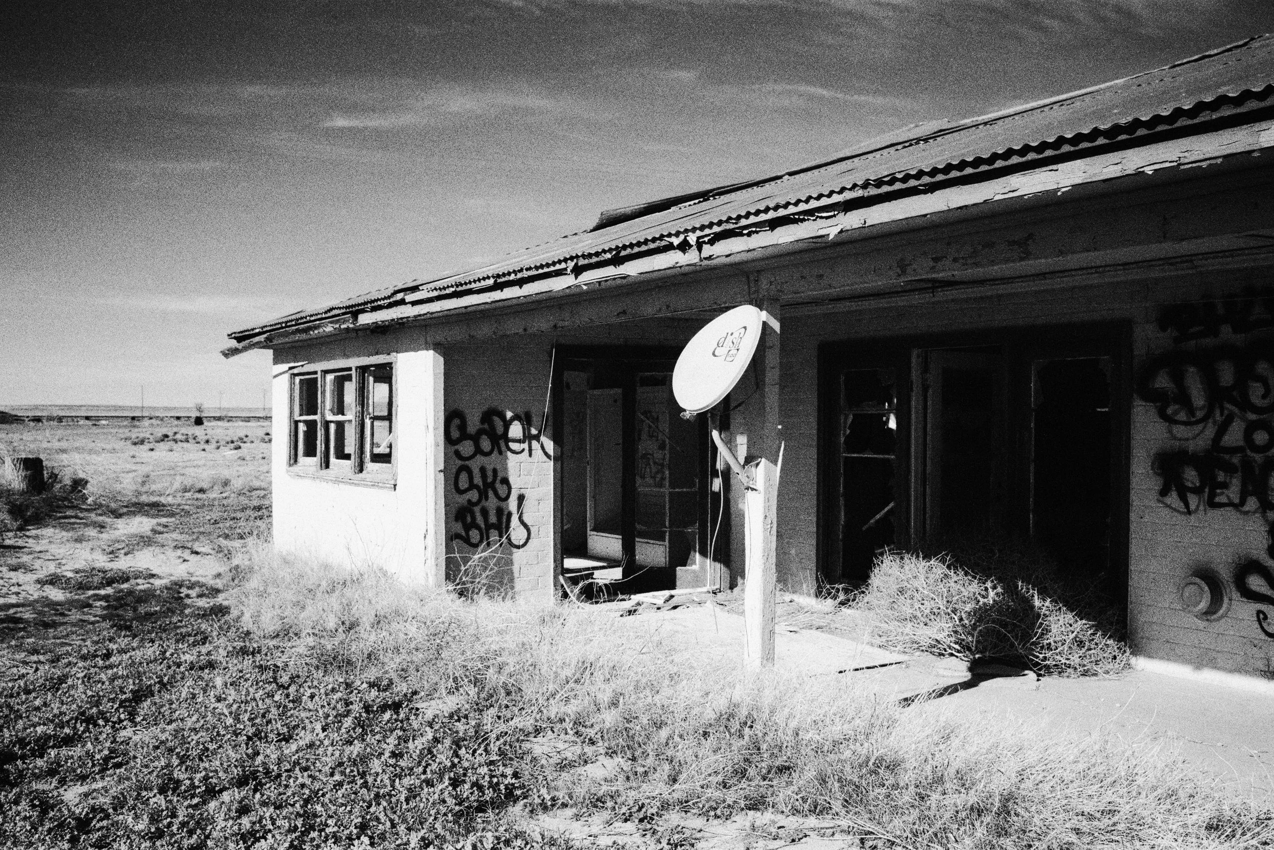 An abandoned, dilapidated house with broken windows and graffiti on the walls, situated in a desert landscape under a cloudy sky, with a satellite dish mounted on a post near the entrance.
