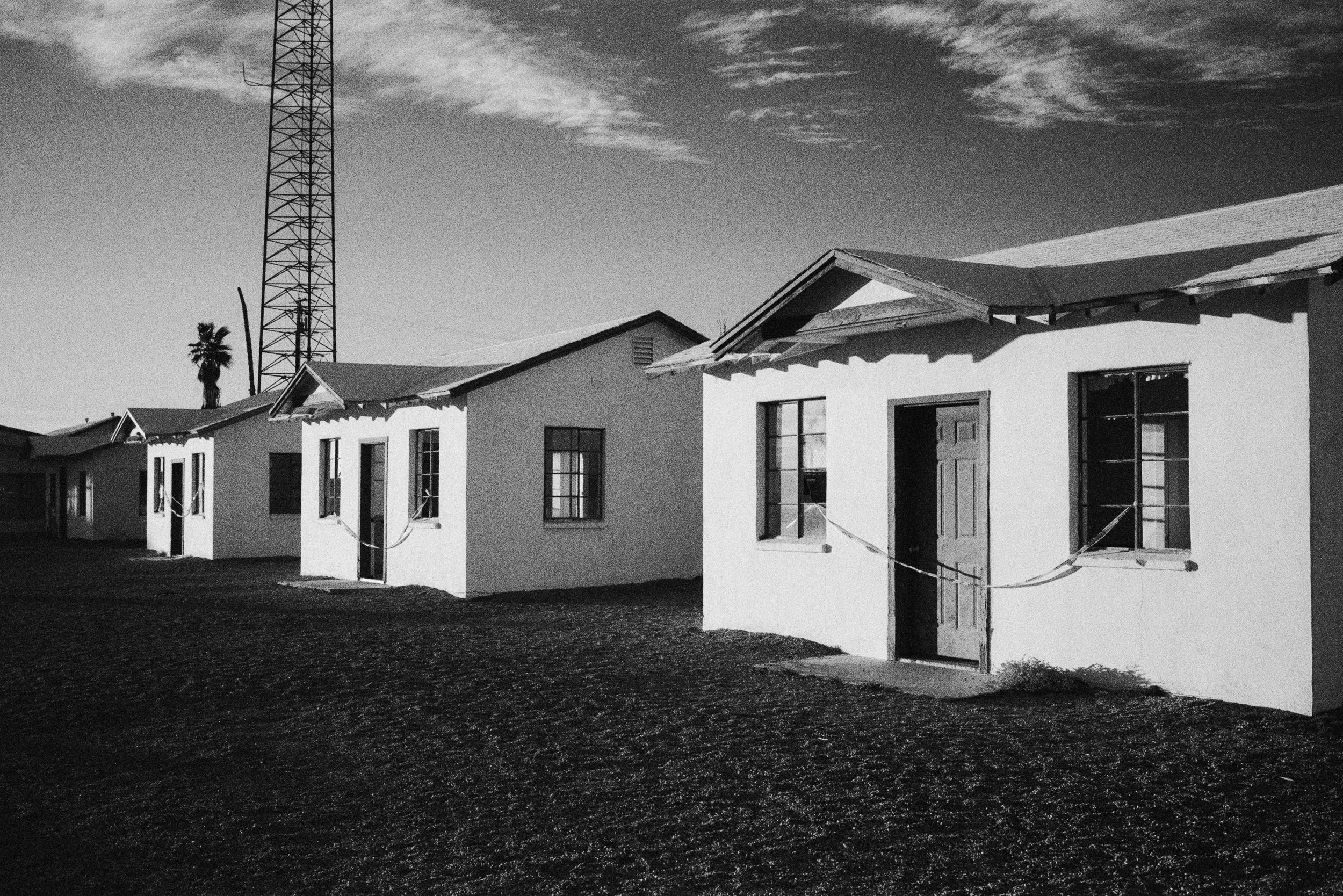 A row of small, white houses with open doors and windows, set against a clear sky with a few clouds. A tall radio tower is visible in the background.