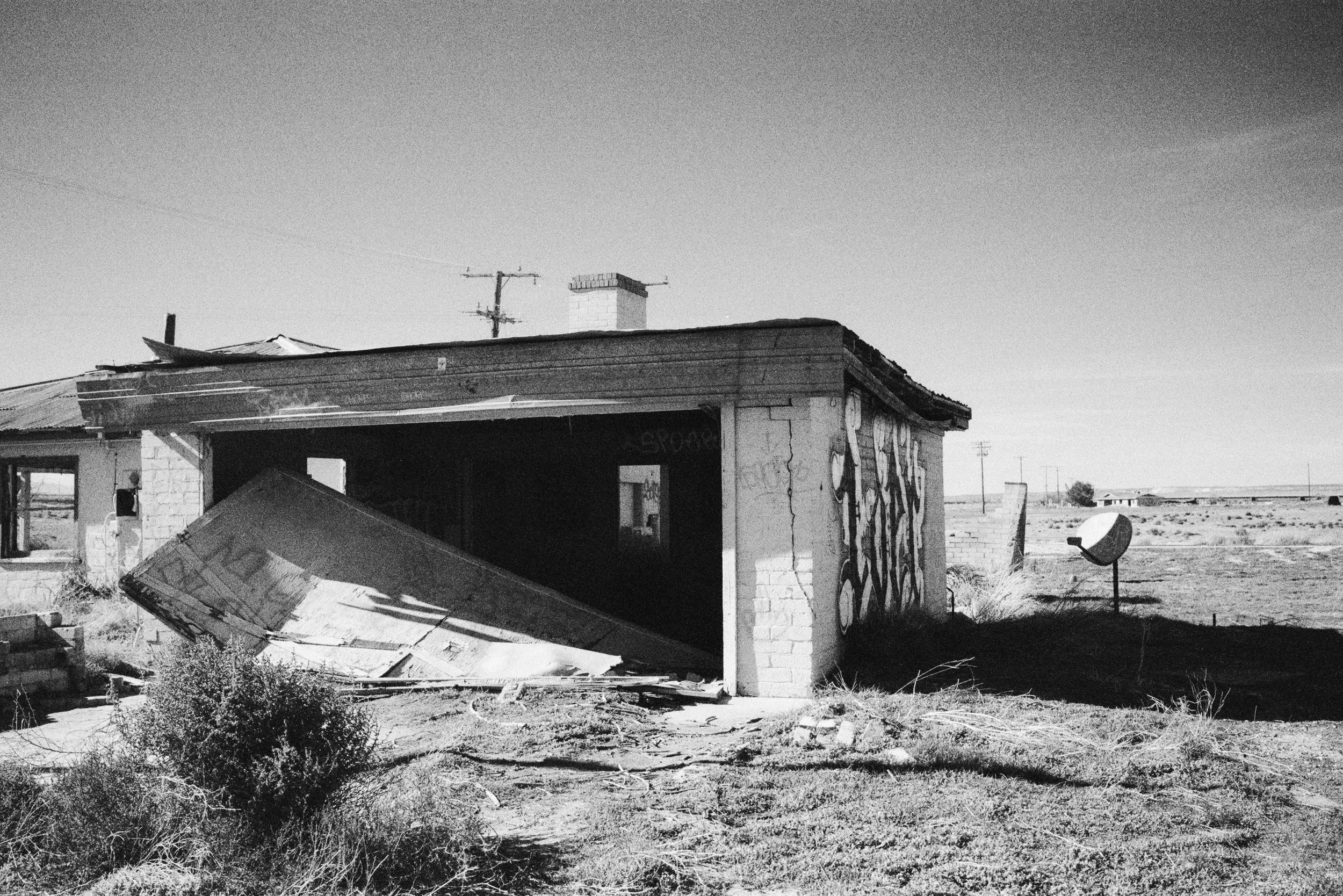 A dilapidated house with a partially collapsed roof and graffiti on the wall, situated in a barren, desert-like landscape with power lines in the background.