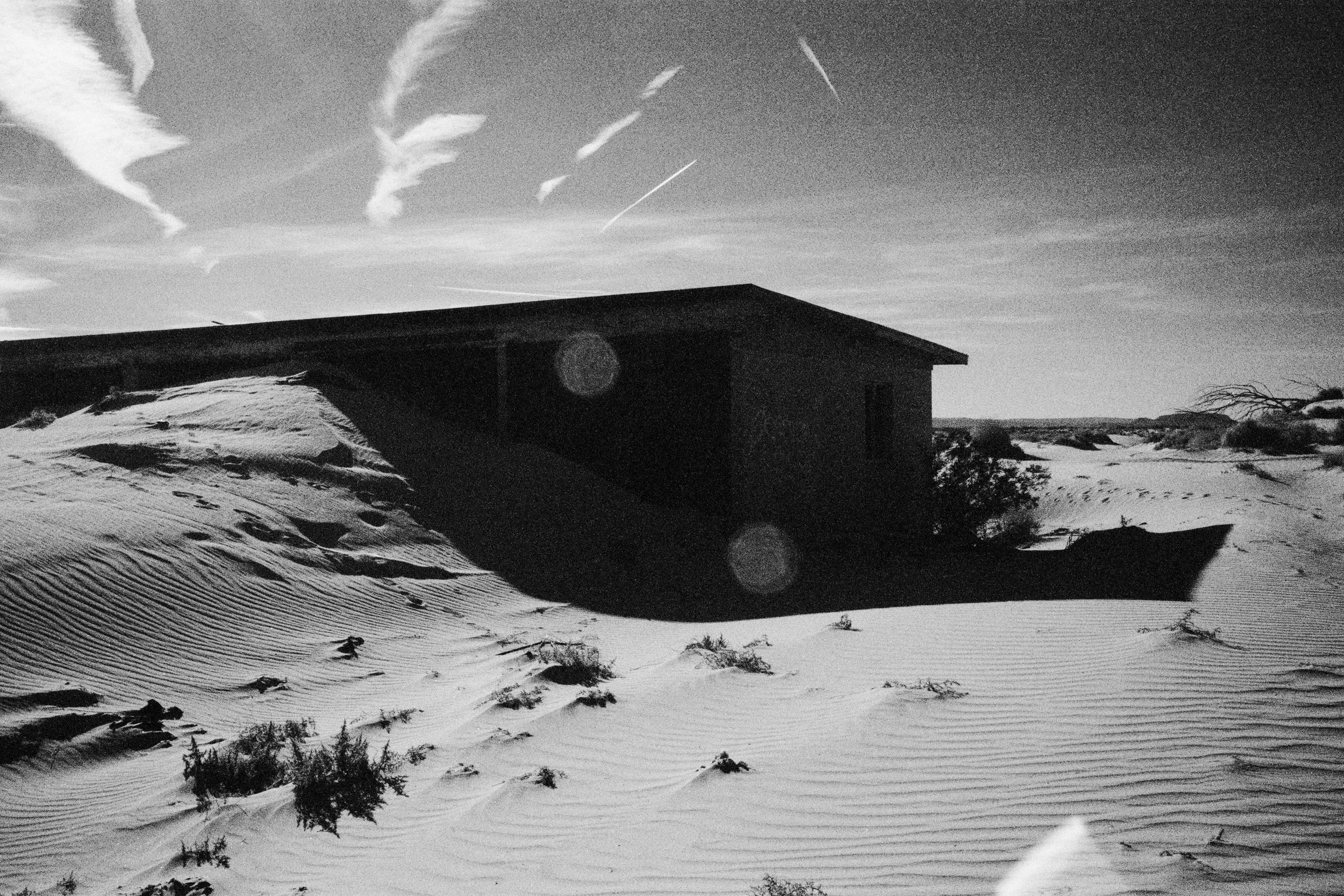 A house in a sand dune landscape with a clear sky and some clouds above, black and white photo.