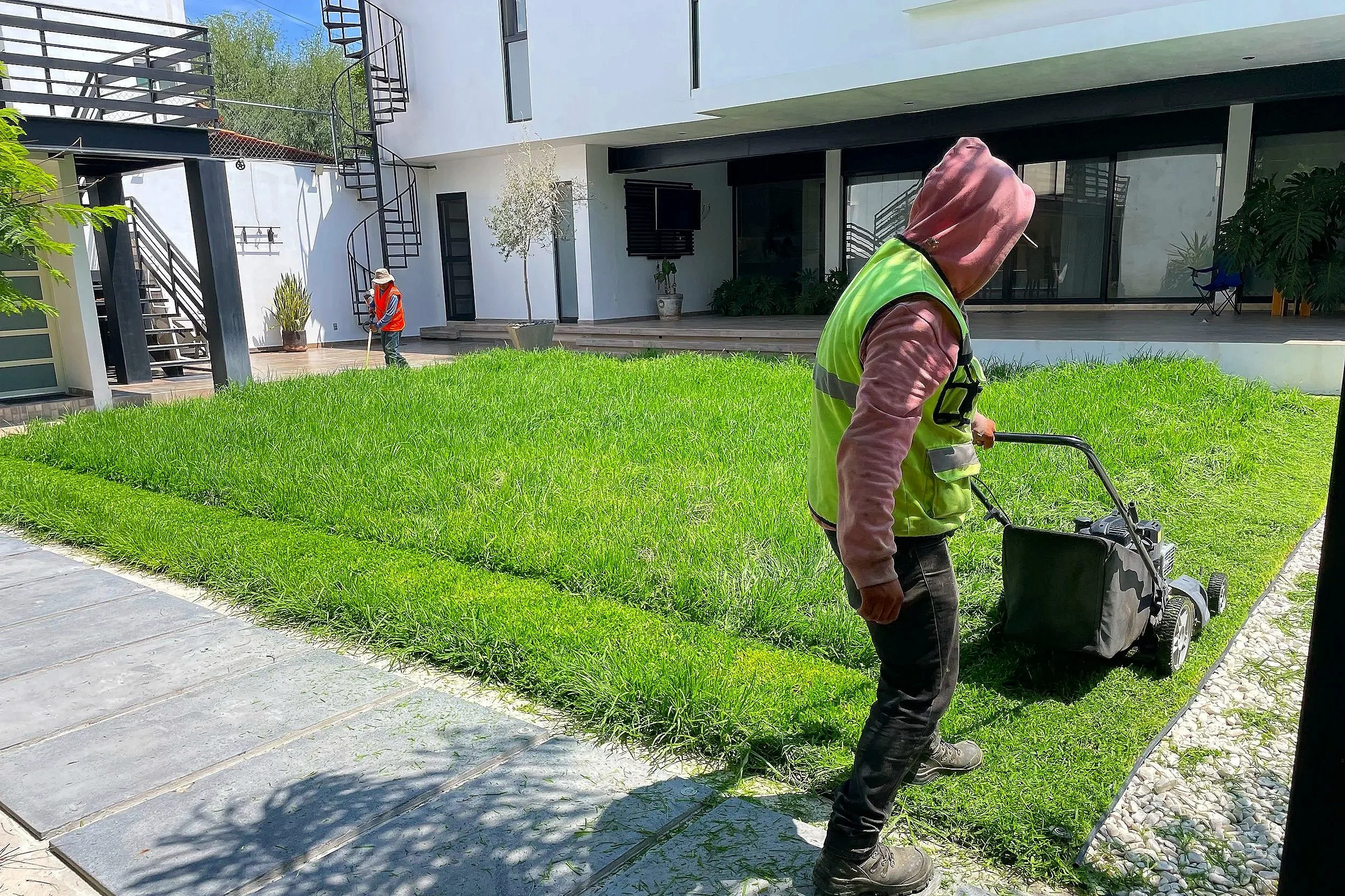 Dos personas trabajando en un jardín en un área residencial moderna, una con cortadora de césped y otra con una rastrillo cerca de una casa blanca y detalles en negro.