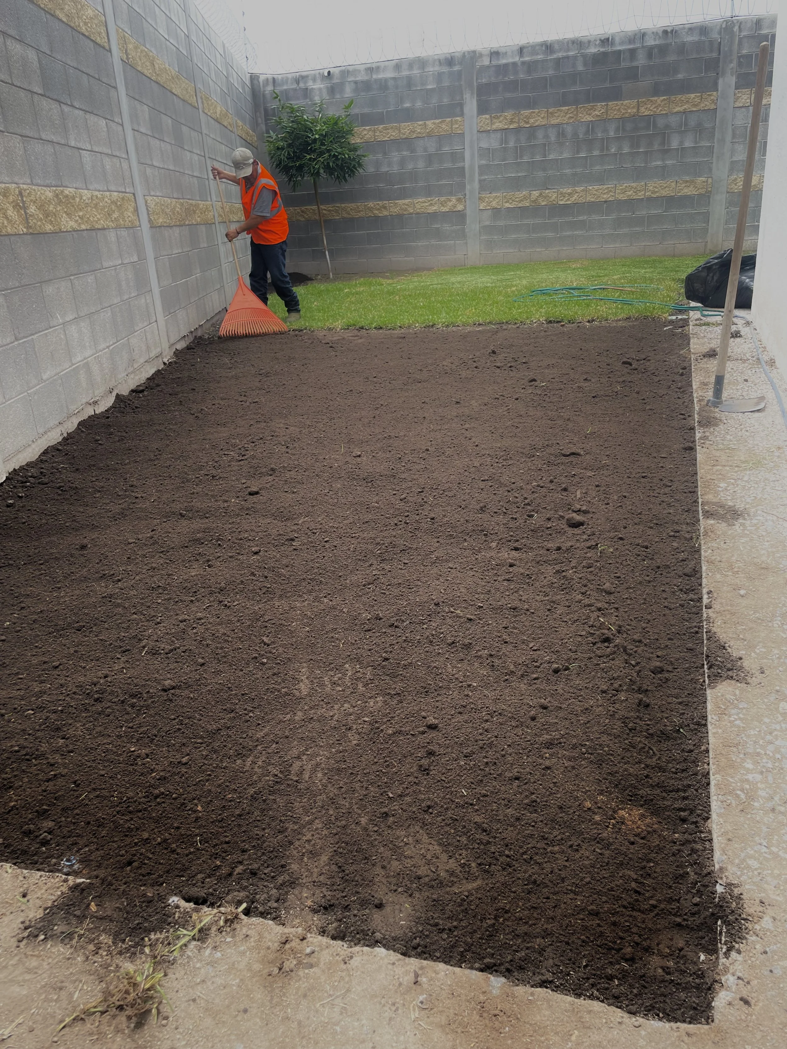 Persona arreglando un jardín con rastrillo en un área de tierra preparada para plantar. Está usando una camiseta gris, un chaleco de seguridad naranja y un sombrero. Hay una cerca de bloques de cemento y césped con un árbol al fondo.