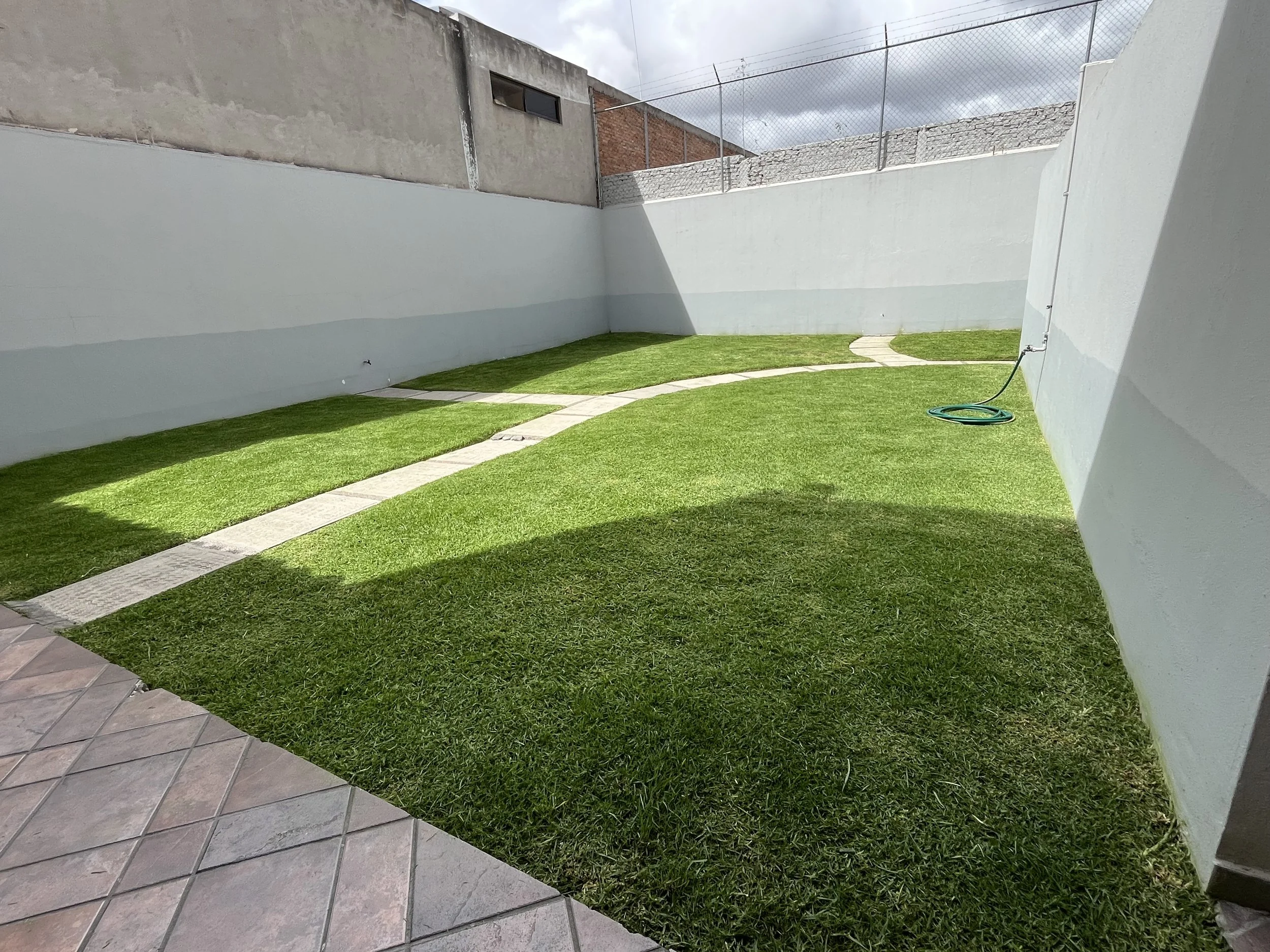 Jardín con césped verde, senderos de piedra, y muro alto gris