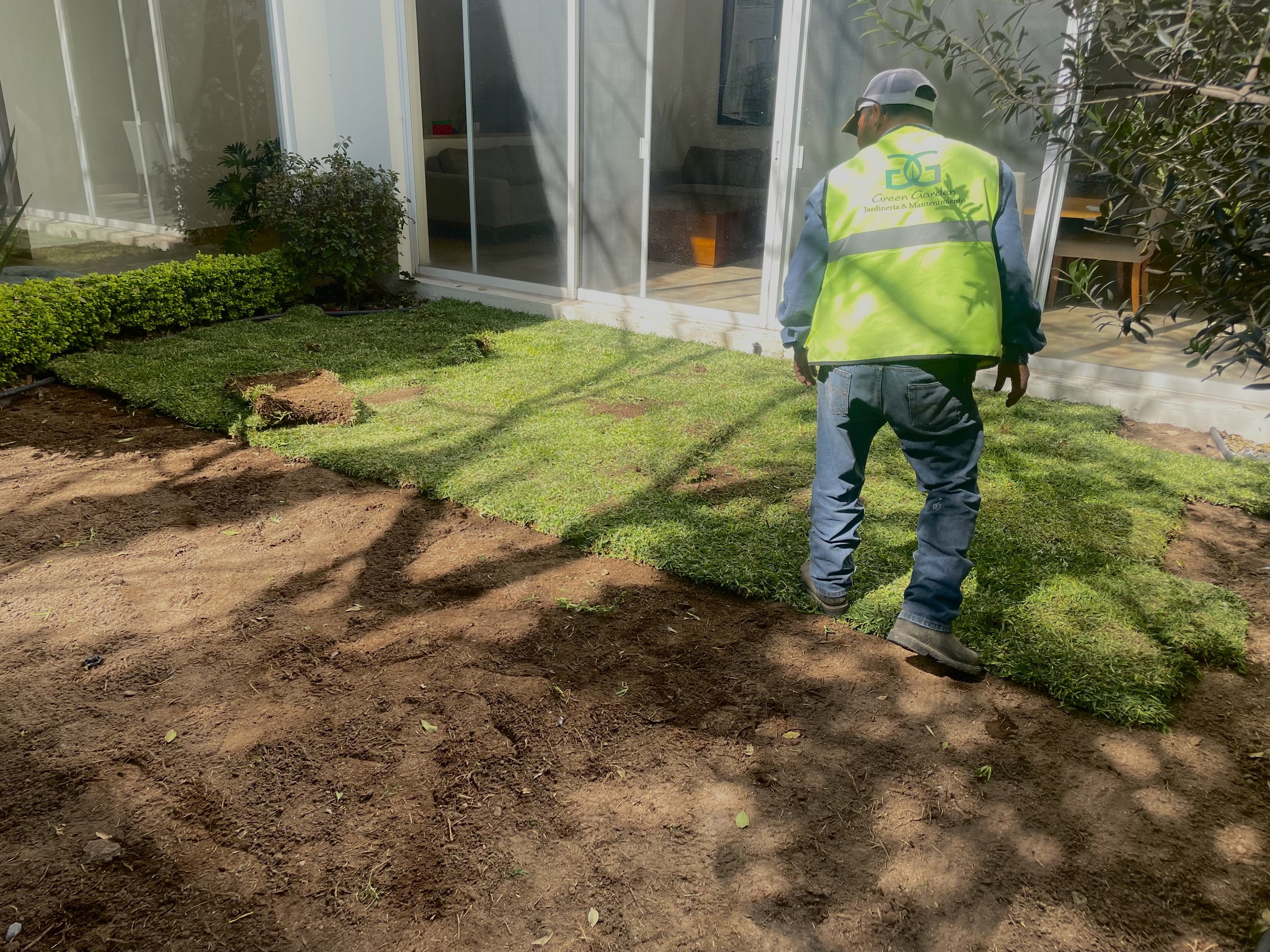Un trabajador realizando el reemplazo de césped en un jardín de una casa, con una vestimenta de seguridad color amarillo y un chaleco que dice 'Green Garden'.
