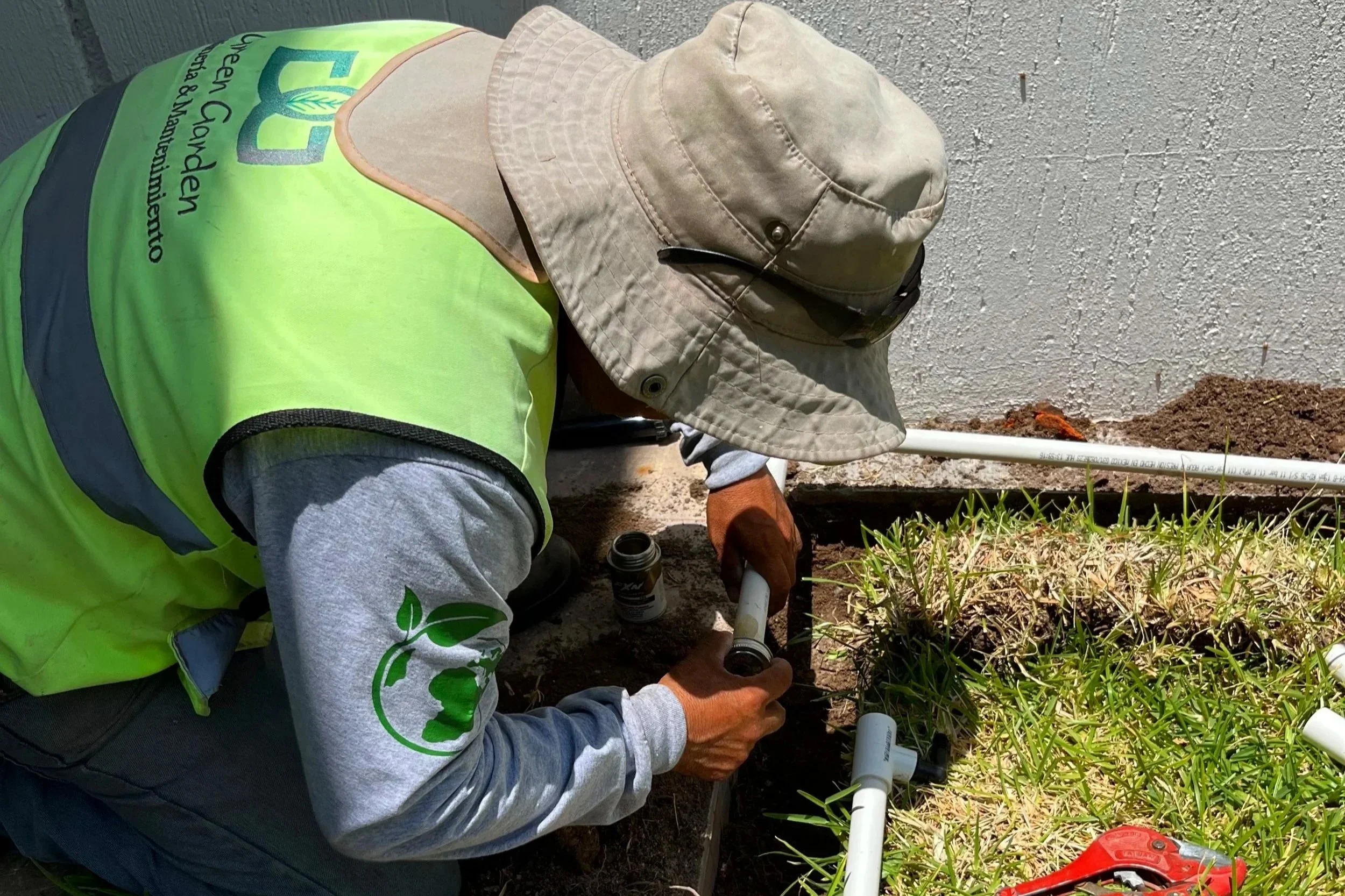 Persona trabajando en la instalación de tuberías de plomería al lado de una pared de cemento, usando gorra, guantes y chaleco de seguridad durante el día.