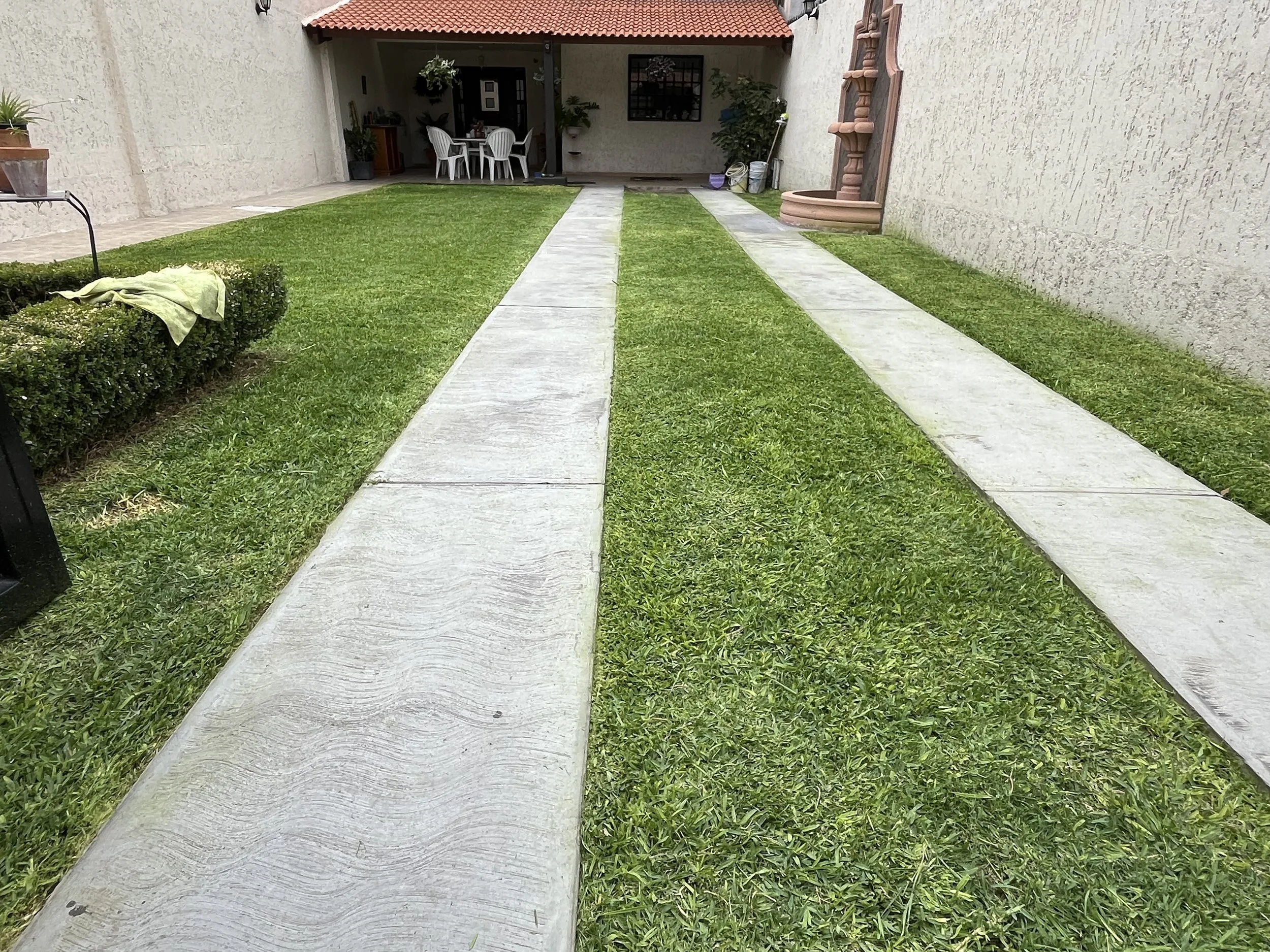 Jardín con césped y camino de concreto que lleva a una zona de comedor con sillas blancas y una mesa, rodeado de paredes de piedra y decoraciones de barro.