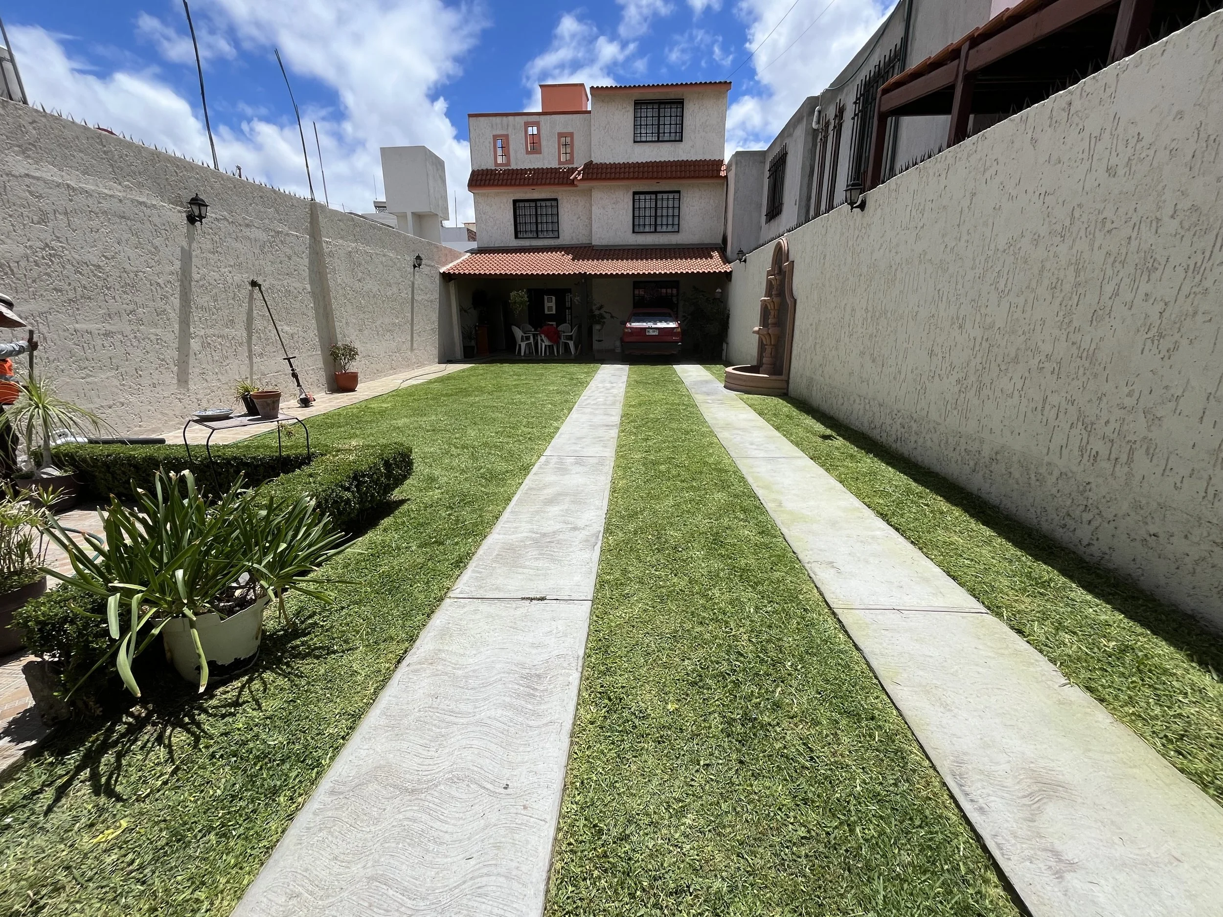 Jardín con césped, caminos de concreto, macetas con plantas, muro alto, casa de varias plantas y coche aparcado en la entrada
