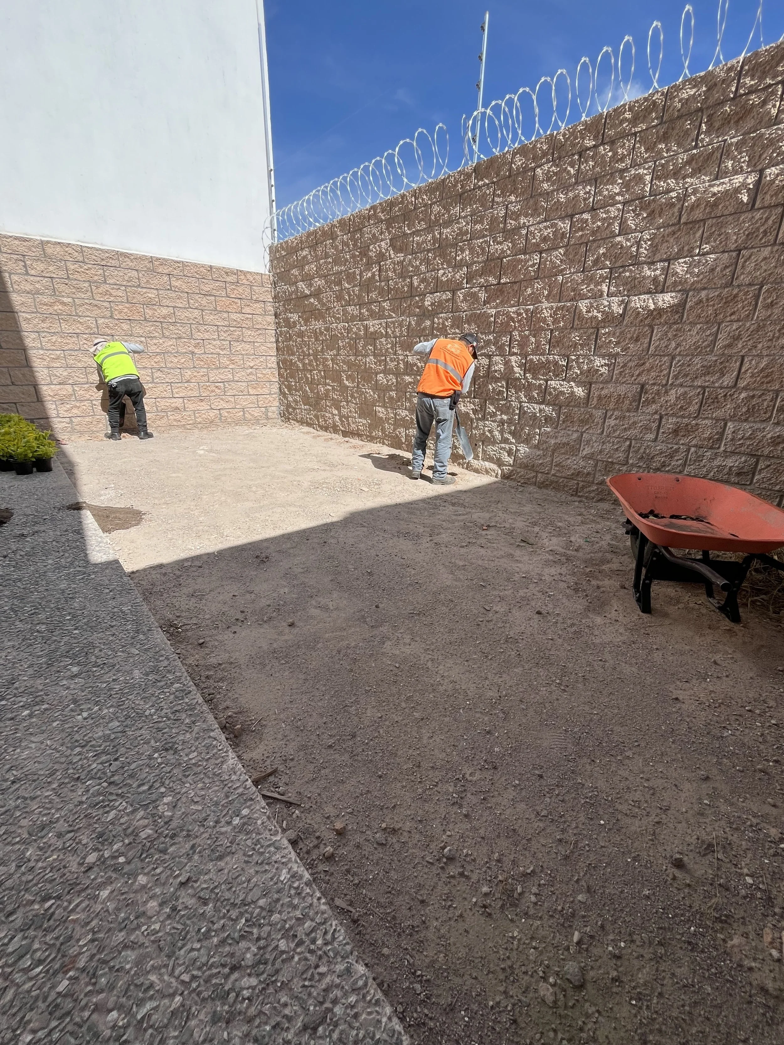 Dos personas trabajando en la construcción de una pared de bloques de piedra, una con chaleco naranja y otra con chaleco amarillo, en un espacio exterior.