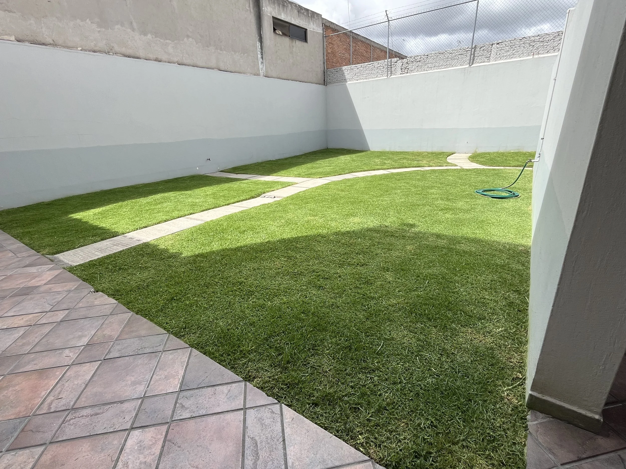 Jardín con césped bien cuidado y camino de piedra, con muro de ladrillos y vivienda cercana, ambiente soleado con algunas nubes.