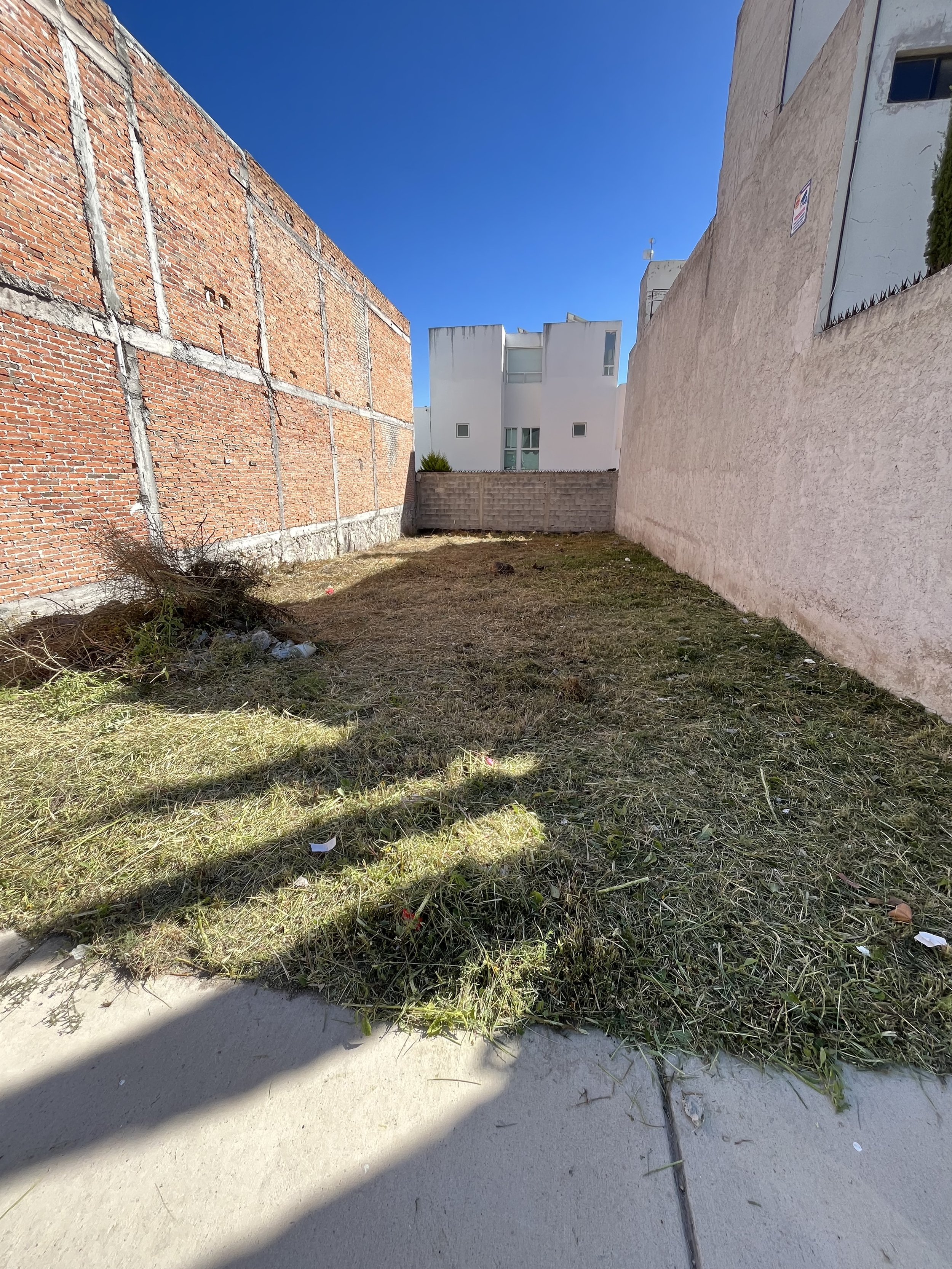 Terreno baldío con césped seco, rodeado por muros de ladrillo y concreto, con edificio blanco al fondo, bajo un cielo despejado.