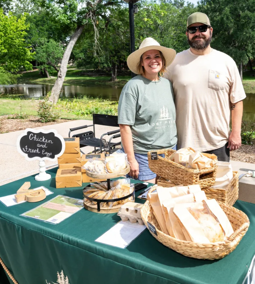Two people smiling at an outdoor market stand selling chicken and fresh eggs, with baskets and a chalkboard sign, in a park setting.