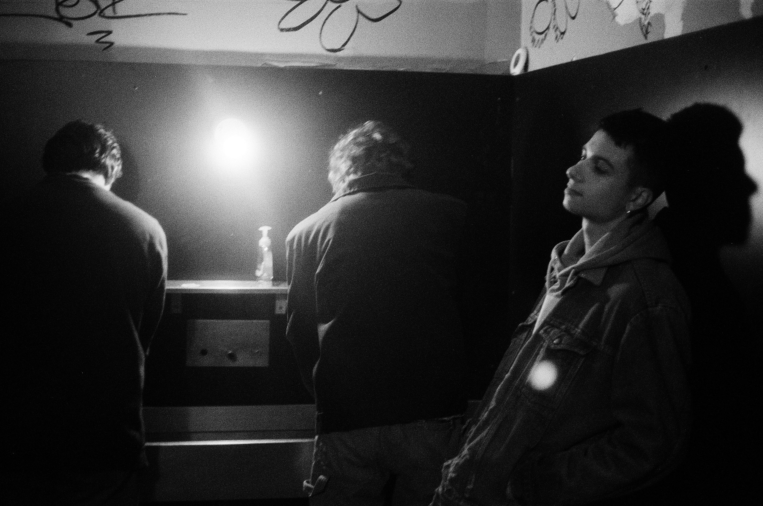 Three people in a dimly lit room, two with their backs turned and one woman facing slightly to the side, standing against a dark wall with floral patterns, near a foosball table, with a bright light illuminating the scene.