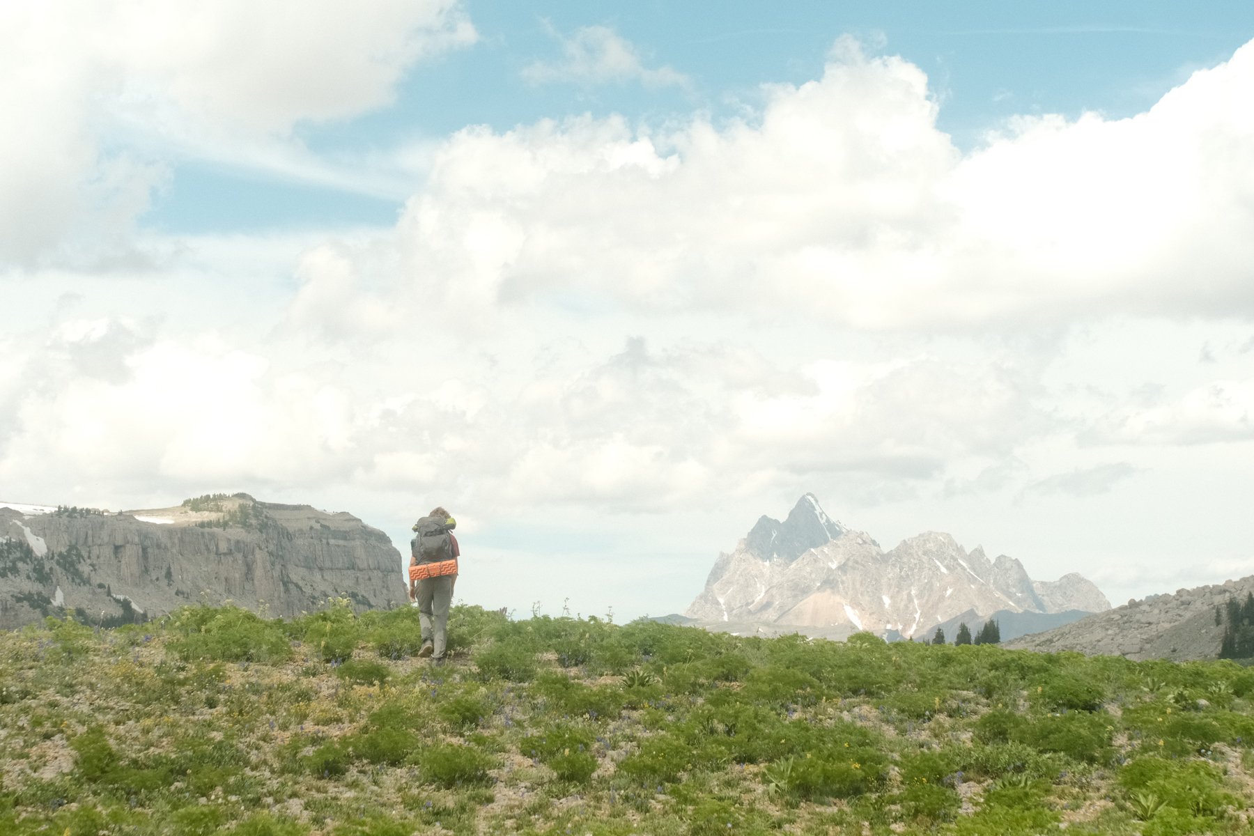 A person hiking in a mountainous landscape with green terrain, rocky cliffs, and mountain peaks in the background under a partly cloudy sky.
