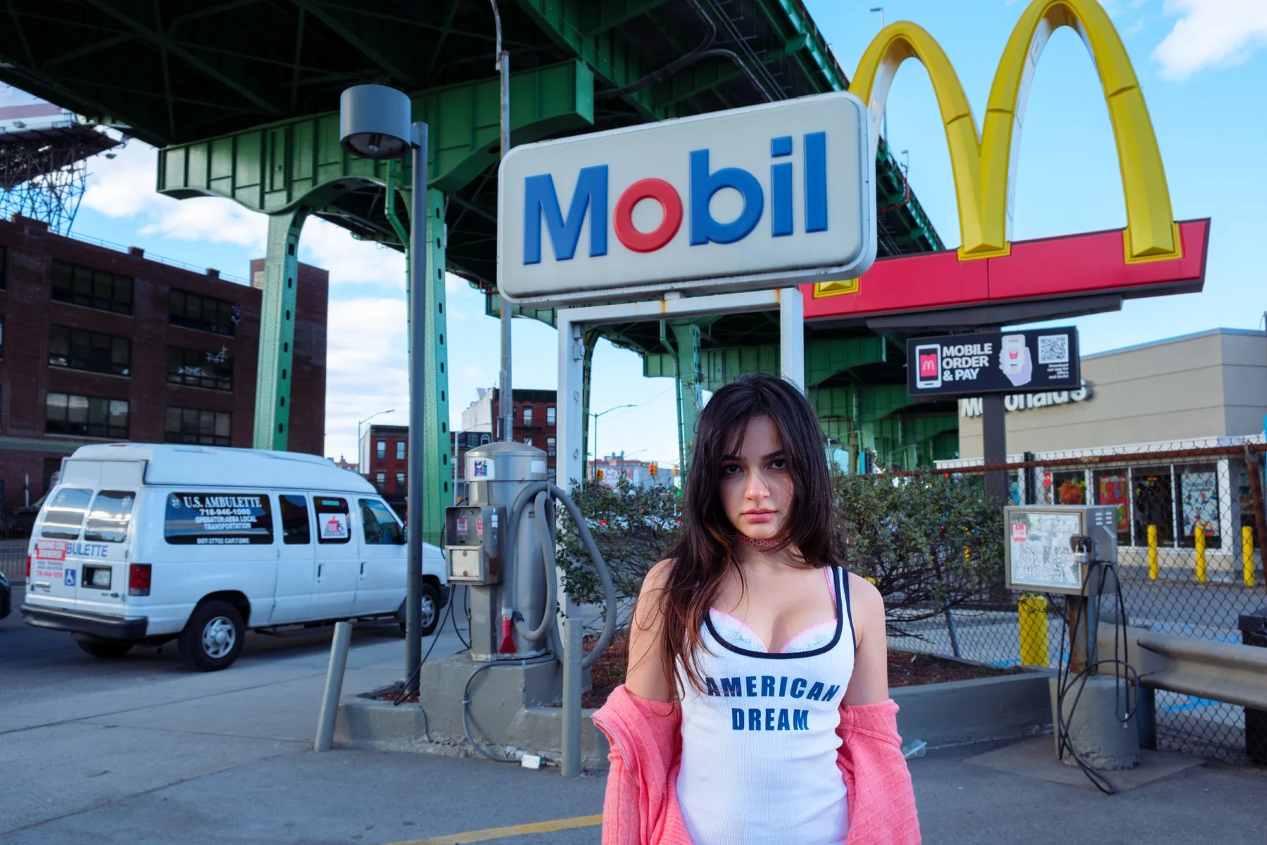A young woman standing in front of a Mobil gas station with a McDonald's sign overhead in an urban setting with a white U.S. Ambulance vehicle parked nearby.
