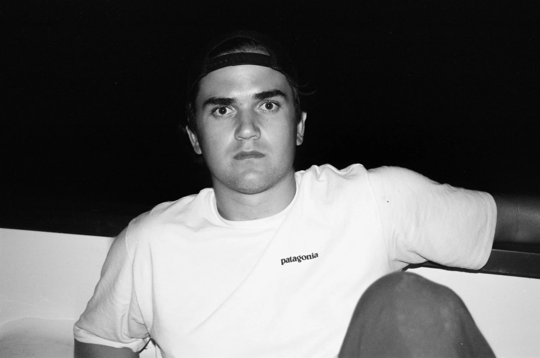A young man with dark hair, wearing a backwards cap and a white Patagonia T-shirt, sitting with his arm resting on a surface, staring directly at the camera, in a black-and-white photo.