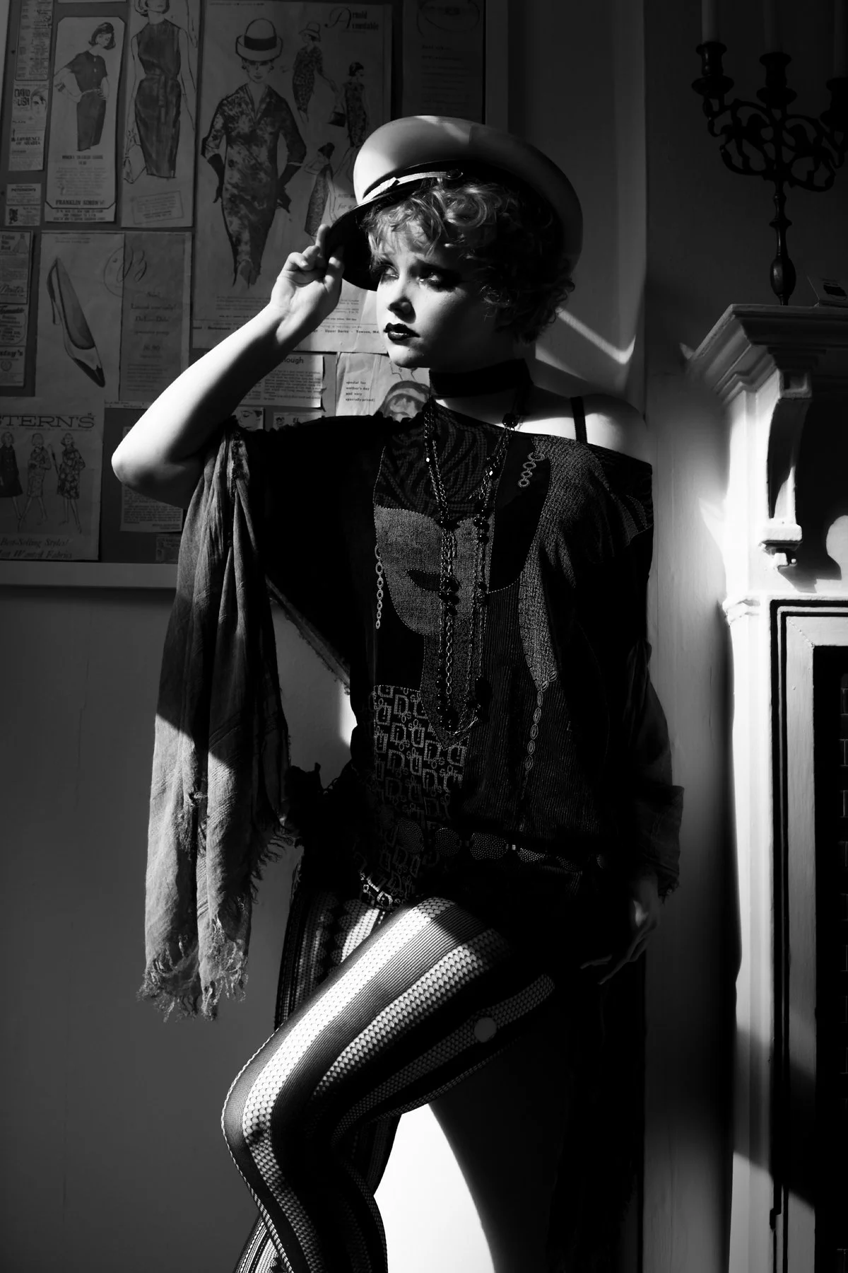 A woman with short curly hair wears a hat, black patterned dress, and patterned tights, standing near a fireplace in a dimly lit room.
