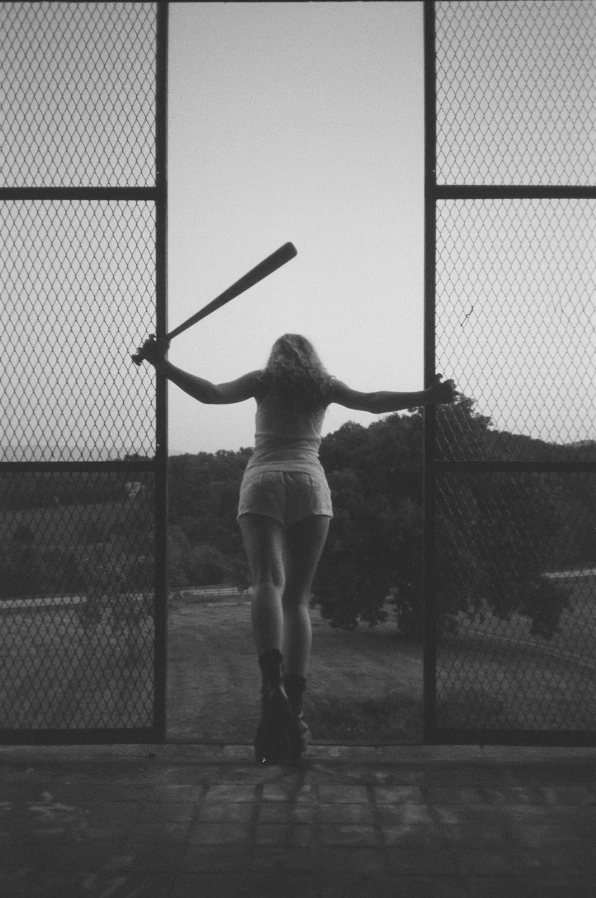 A woman stands with her back to the camera, holding a baseball bat in her right hand, across a chain-link fence, with trees and a hill in the background during twilight.