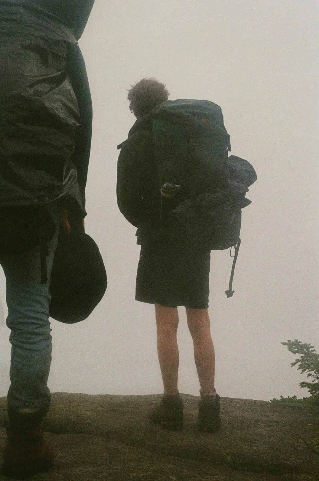 Two hikers with backpacks on a mountain rock in dense fog or clouds, one wearing shorts and the other wearing pants.