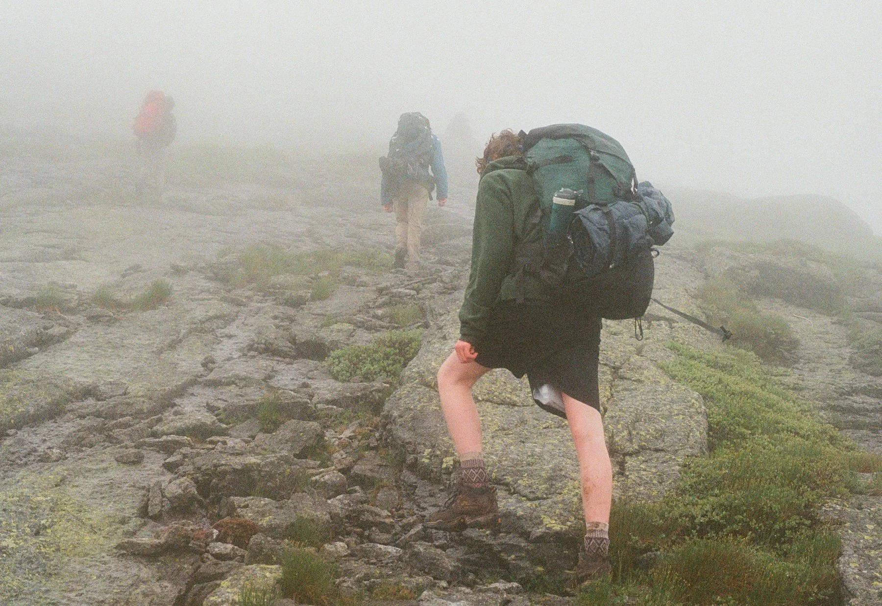 Hikers walking on a rocky trail through dense fog, wearing backpacks and outdoor gear.