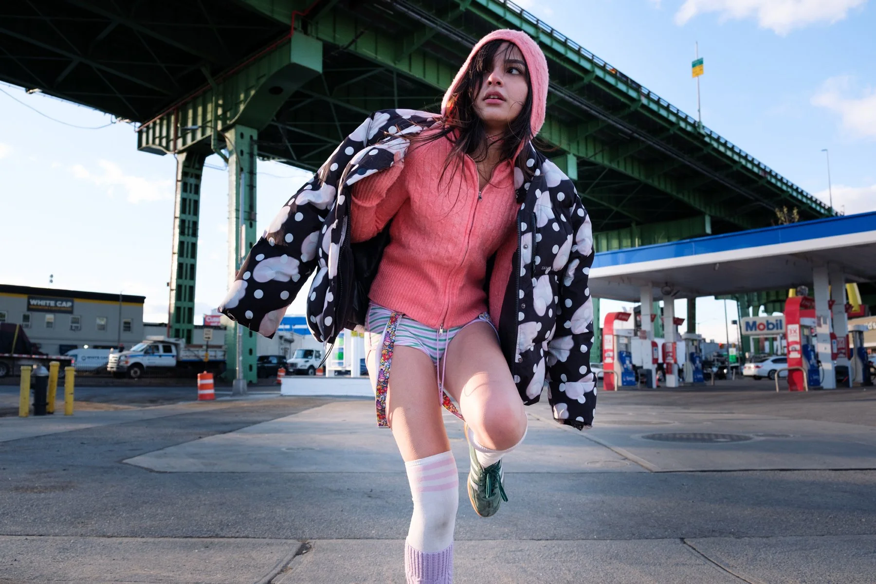 A young woman wearing a pink hooded sweater, a black patterned jacket, striped shorts, and colorful socks, kneels on one knee at a gas station under a bridge, with gas pumps and cars in the background.