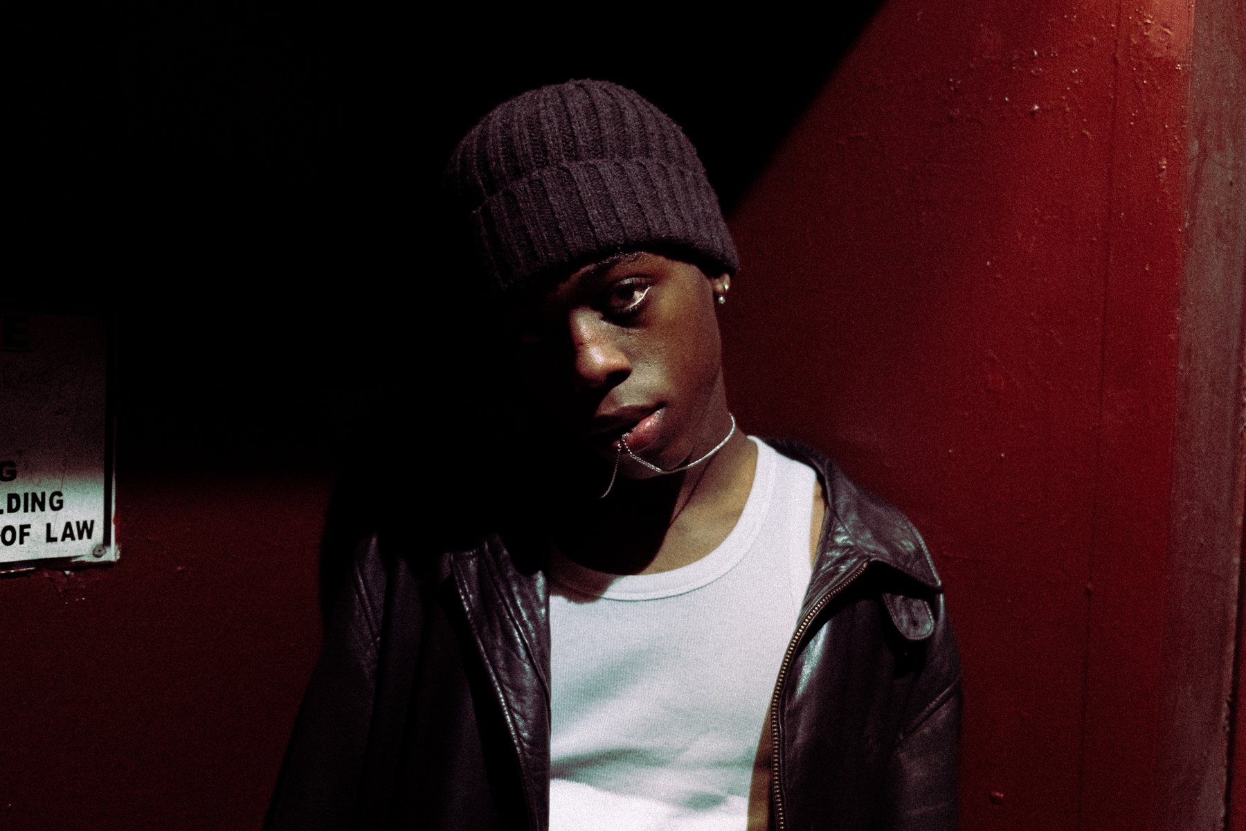 A young person wearing a dark knit beanie, a white shirt, and a black leather jacket, standing against a dark red wall with shadows covering part of their face.