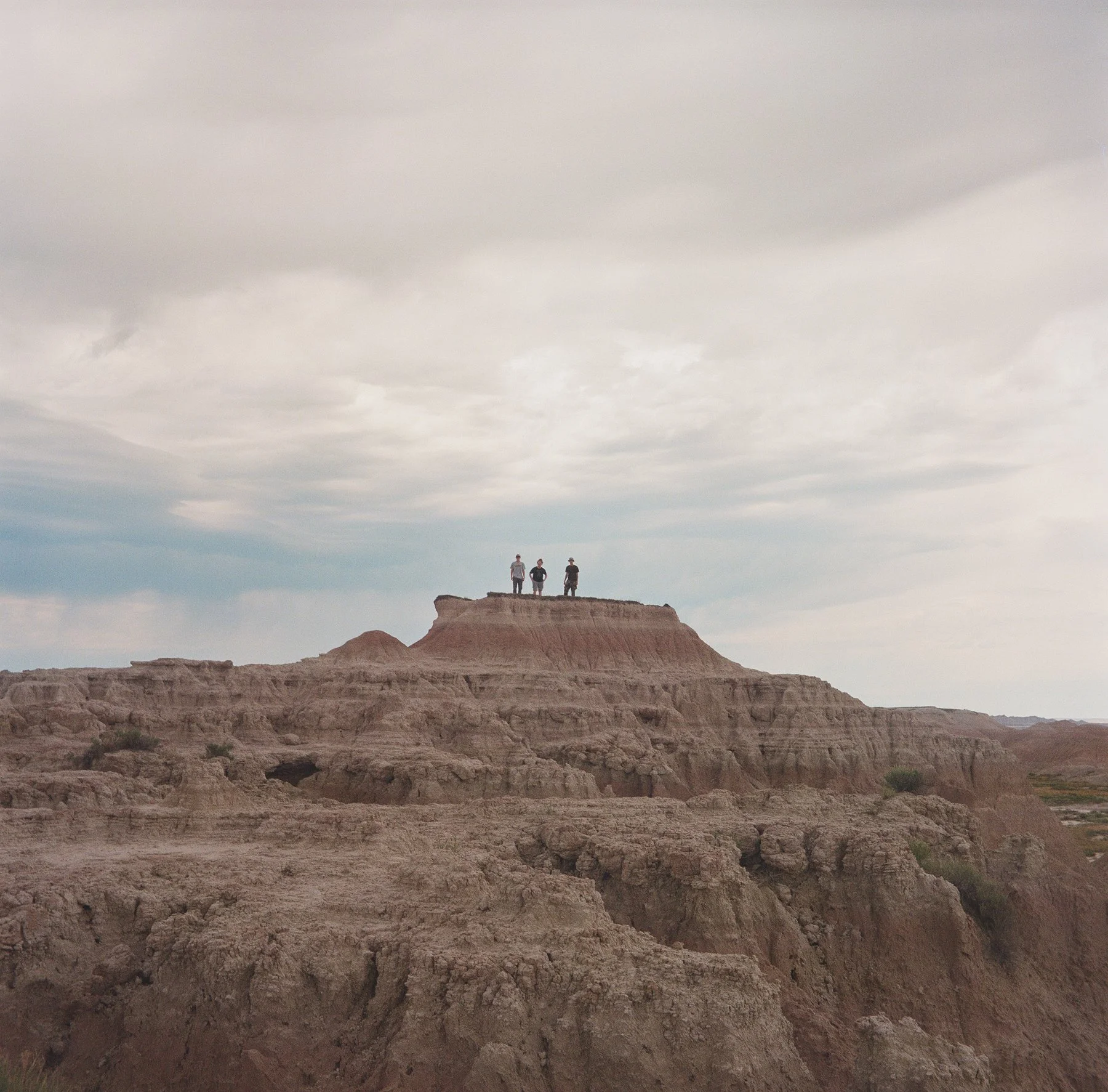 Three people standing on top of a large layered rock formation in a desert landscape, under a cloudy sky.