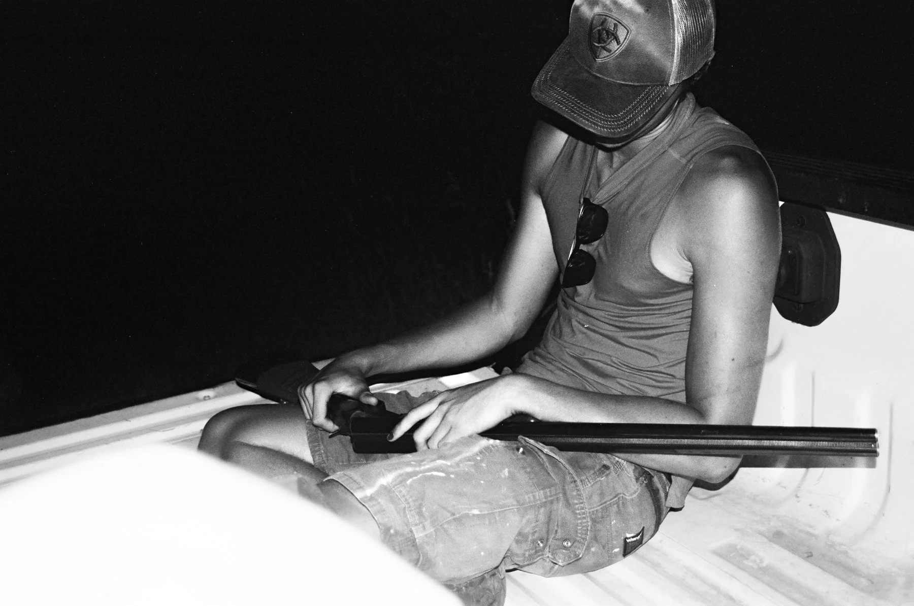 A man sitting in the back of a vehicle, wearing a sleeveless shirt, cap, sunglasses hanging from his shirt, and cargo shorts, holding a rifle.