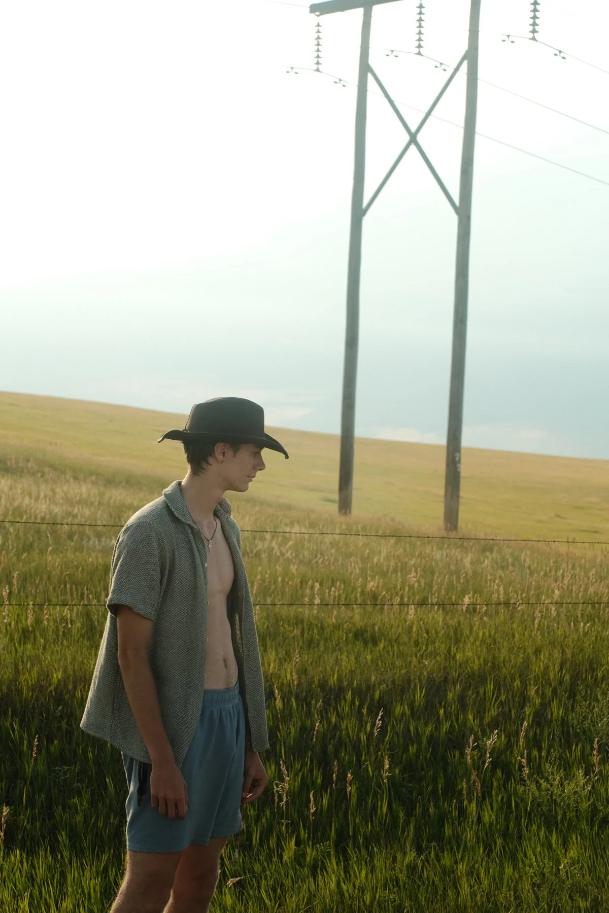 Young man standing in a grassy field wearing a cowboy hat, open shirt, and shorts, with power lines in the background on a cloudy day.