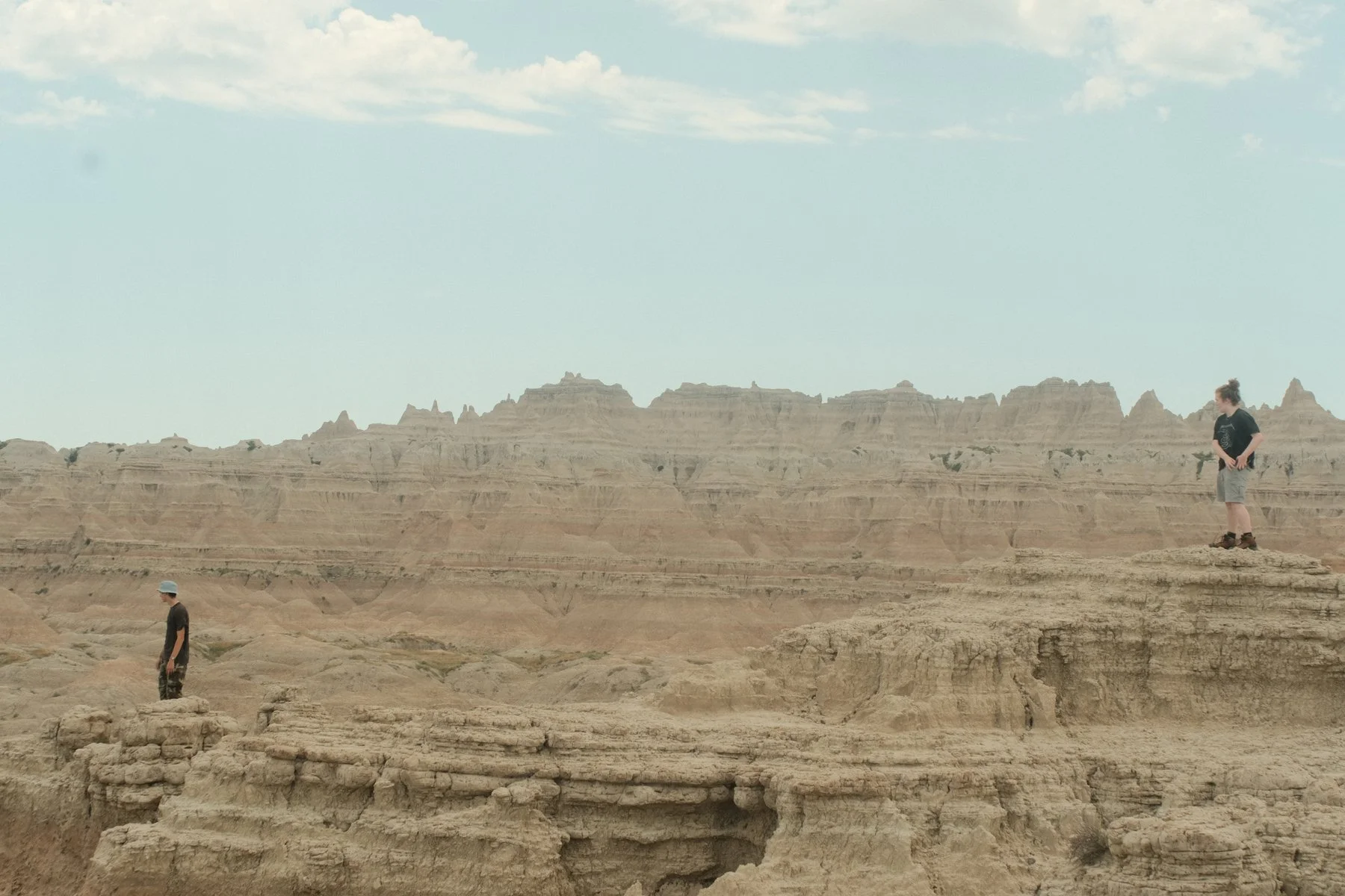 Two people standing on the edge of a canyon with layered rock formations, under a partly cloudy sky.