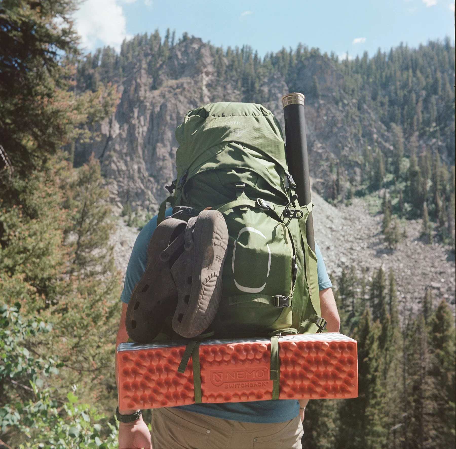 A person hiking in a mountainous forest carrying a large green backpack with a shoe attached to it and a foam sleeping pad strapped to the bottom, with rocky cliffs and pine trees in the background.