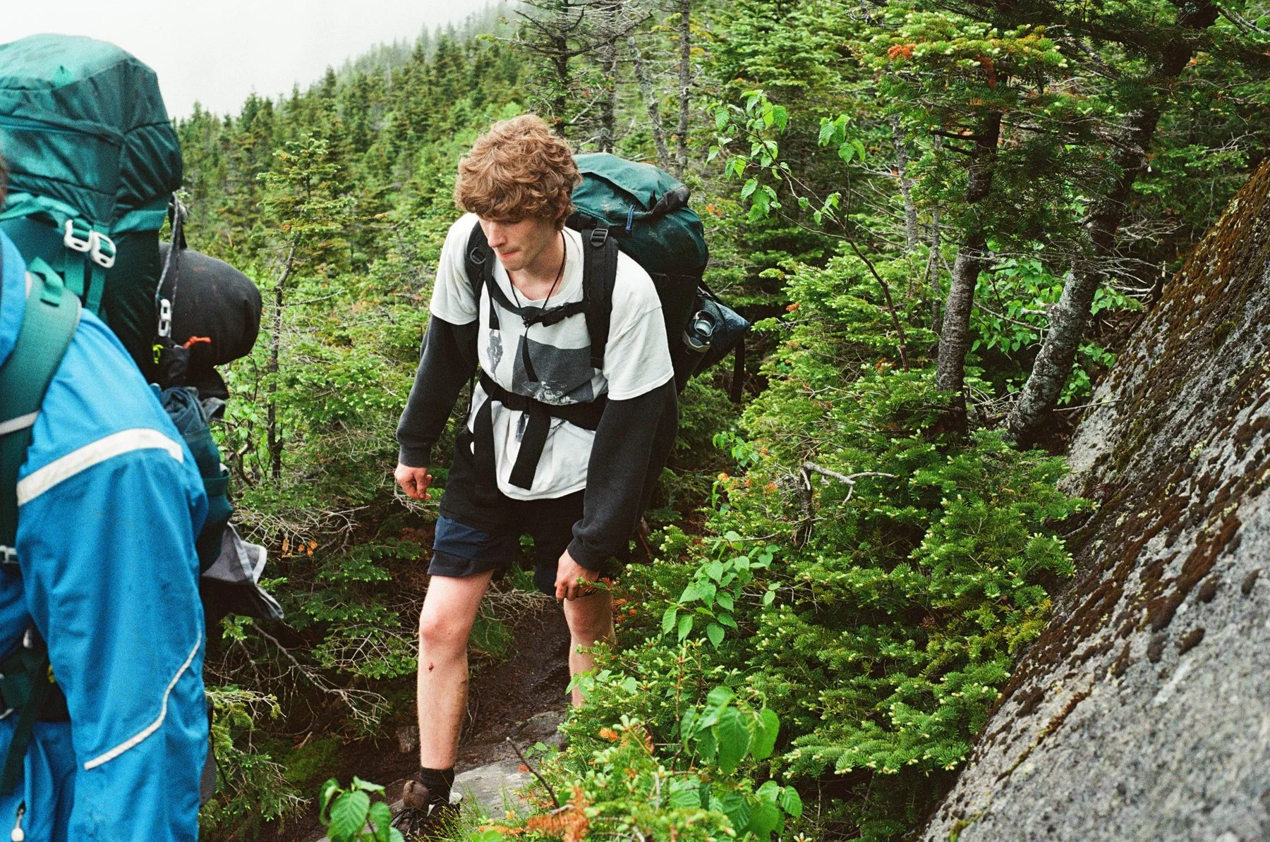 A young man with curly brown hair, wearing a white T-shirt over a black long-sleeve shirt, black shorts, and hiking boots, carrying a large backpack, is descending a steep rocky trail in a forest surrounded by green trees and bushes.