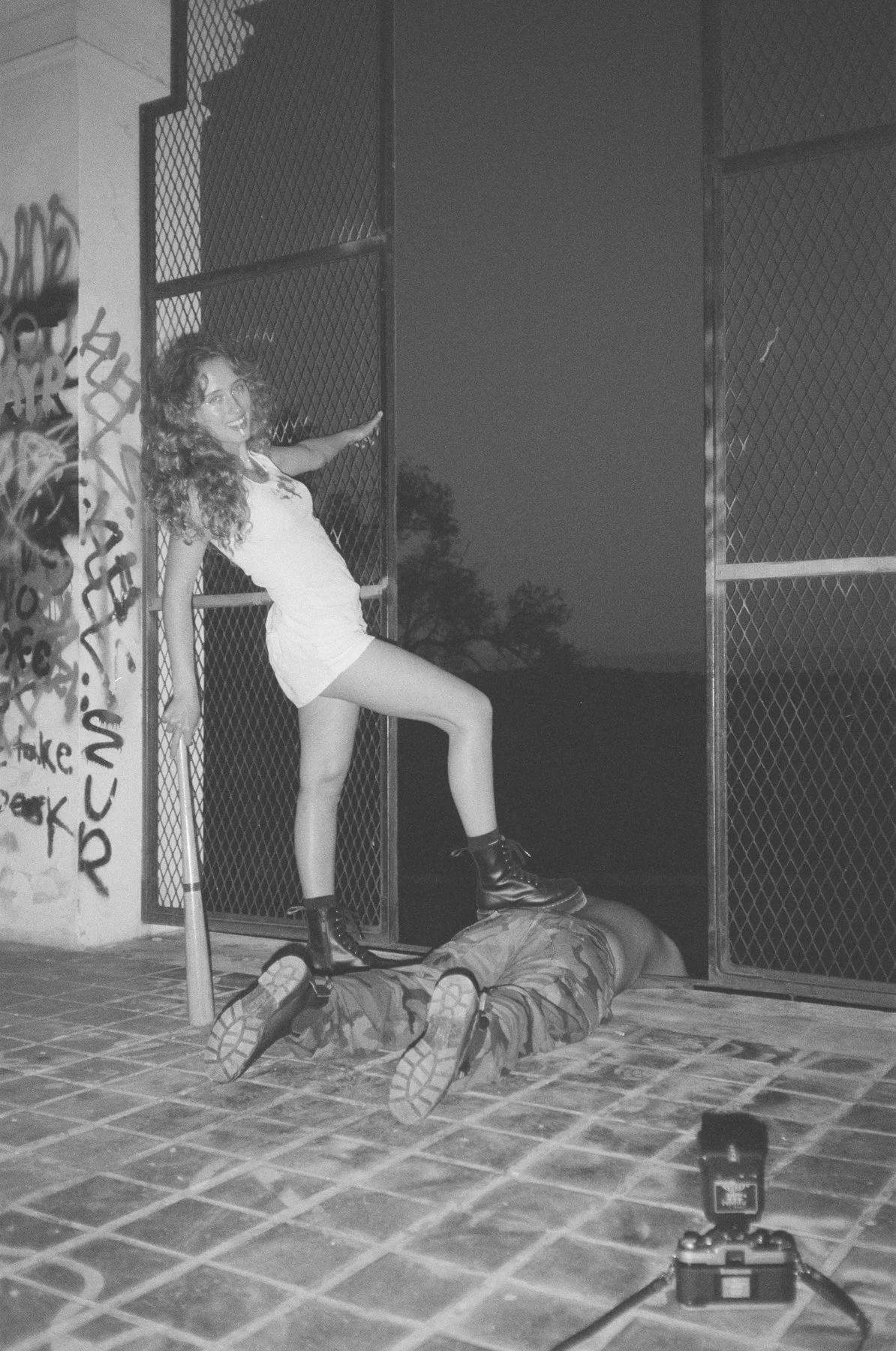 A woman with curly hair stands on a man lying on the ground, examining graffiti-covered walls and an open gate at night.