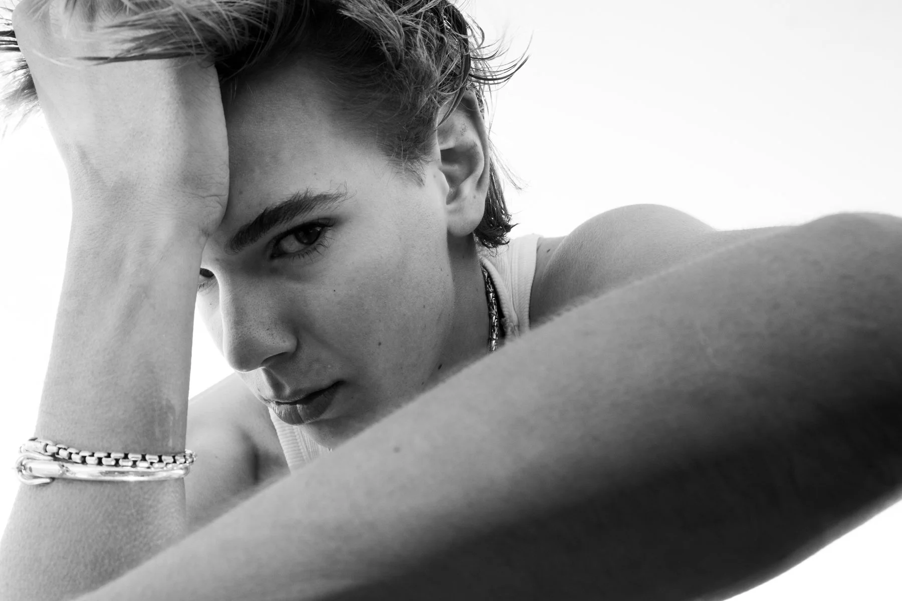 Black and white close-up photograph of a young woman with short hair, gazing into the camera with a serious expression, resting her head on her hand, wearing bracelets and a necklace.