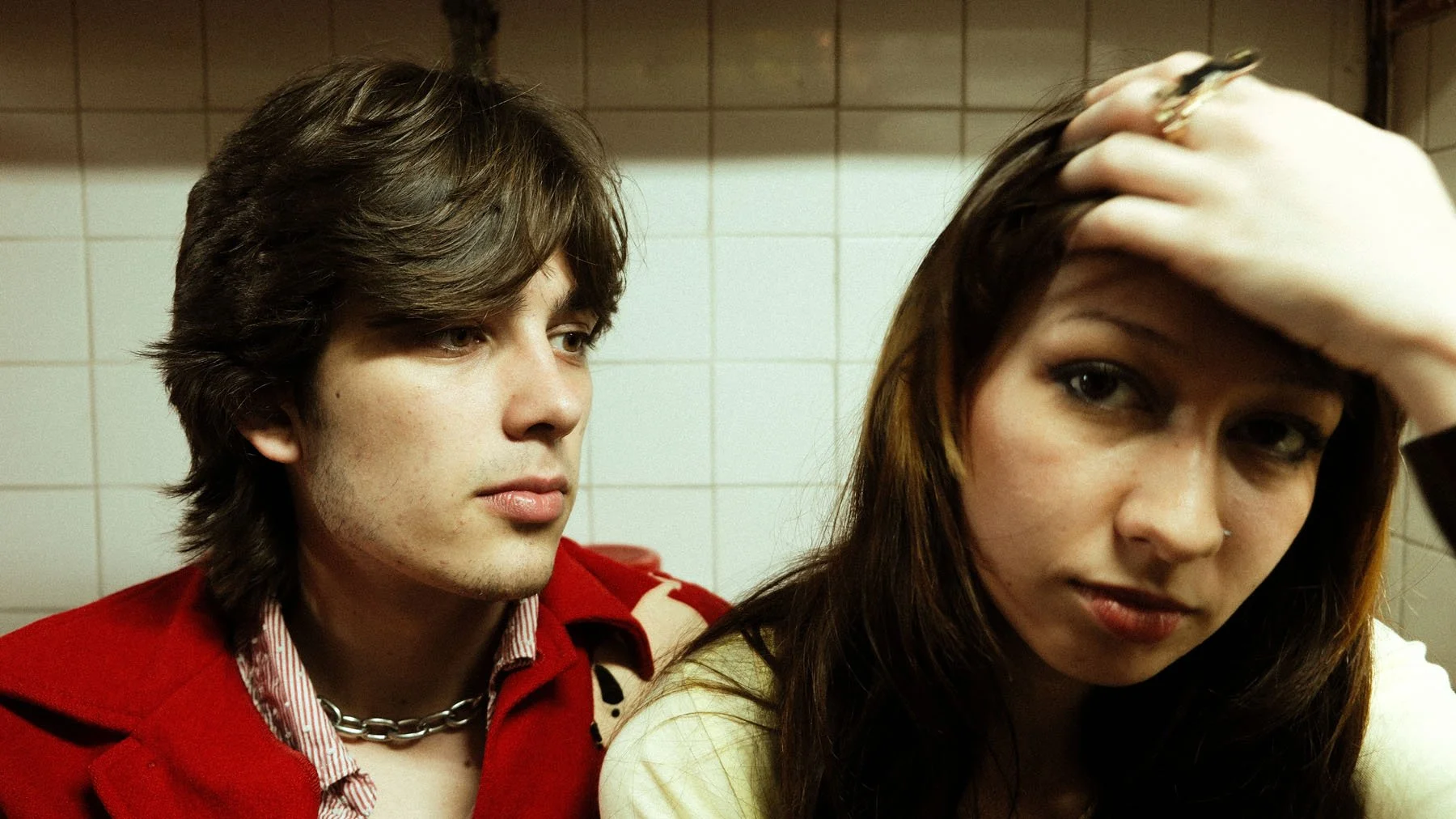 A young man with brown hair and a young woman with dark hair, both with light skin, in a bathroom with tiled walls. The woman is touching her forehead and the man is looking at her.
