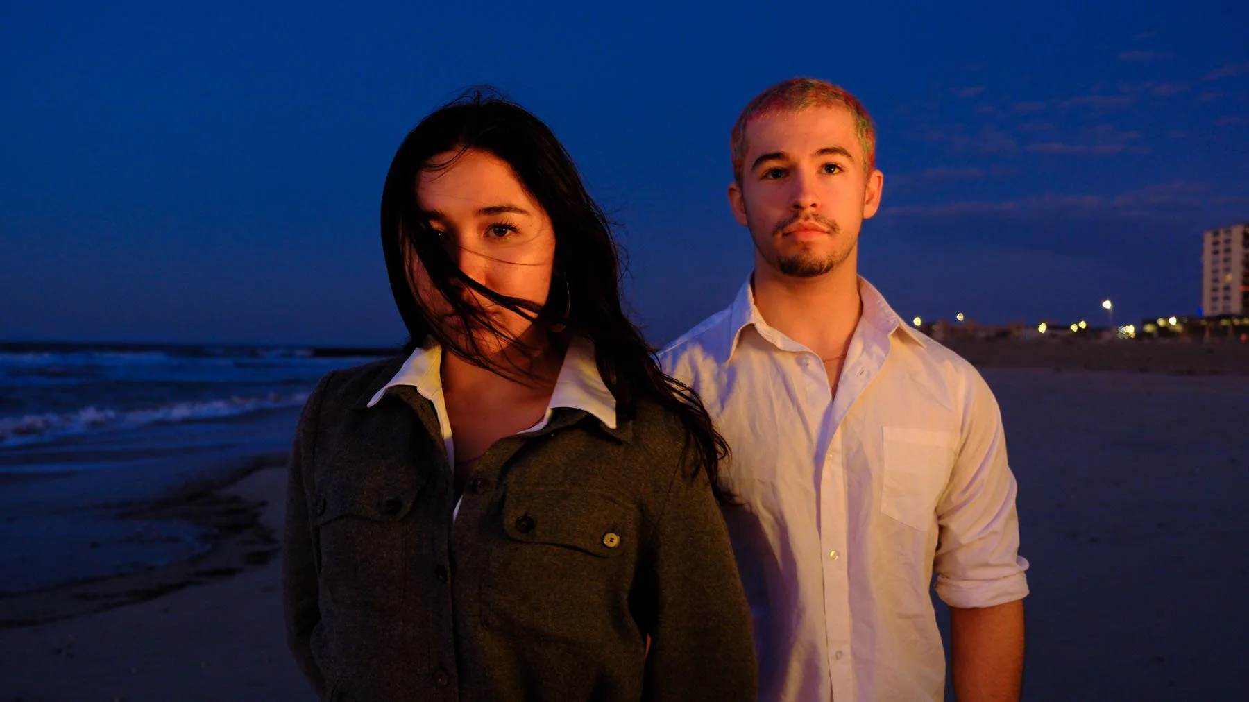 A woman and a man standing on a beach at dusk, with the ocean and city lights in the background.