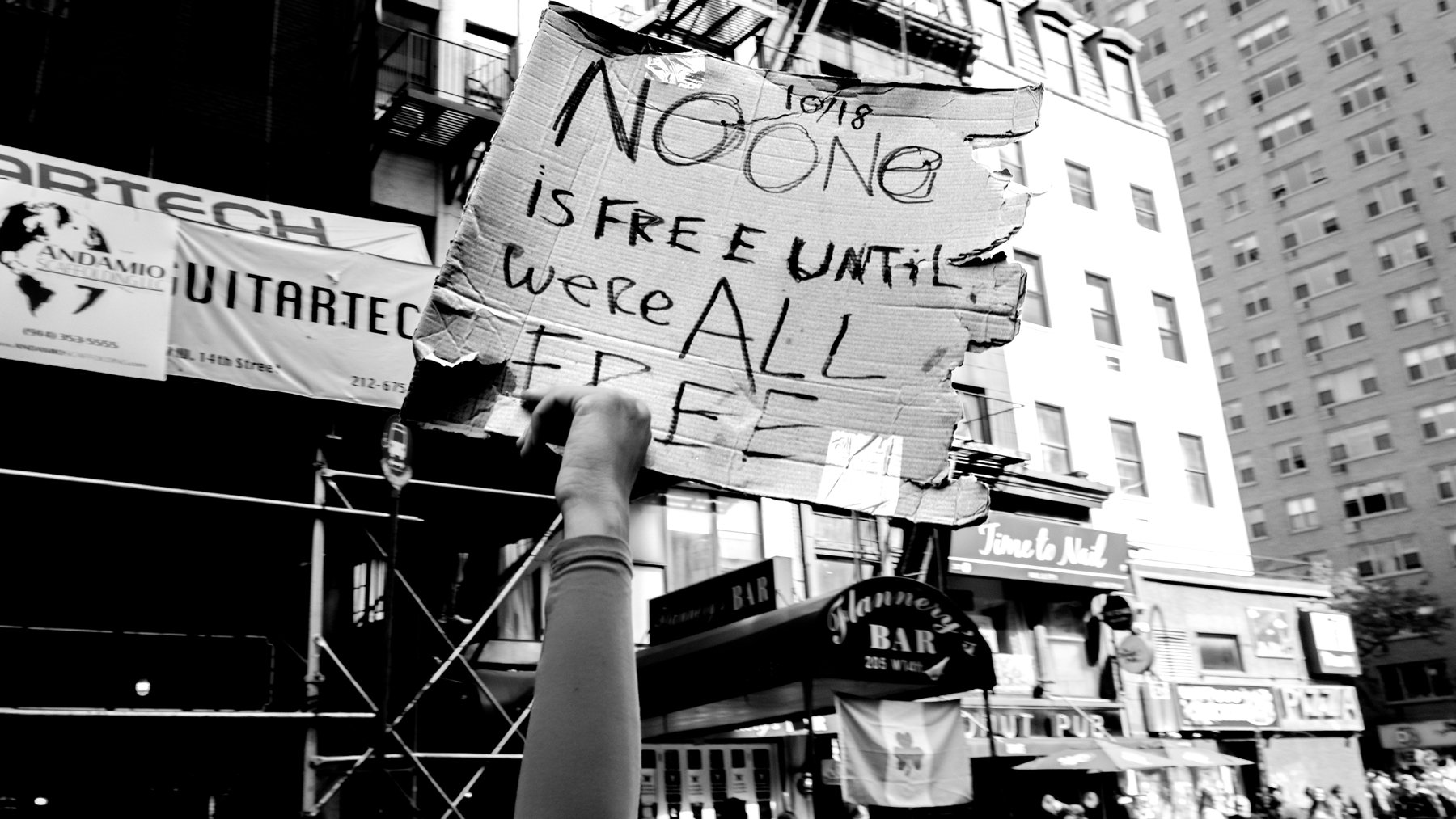 A person holding a cardboard protest sign in an urban street scene with buildings and bars in the background. The sign reads, "NO ONE IS FREE E UNTIL WE ALL ARE FREE."