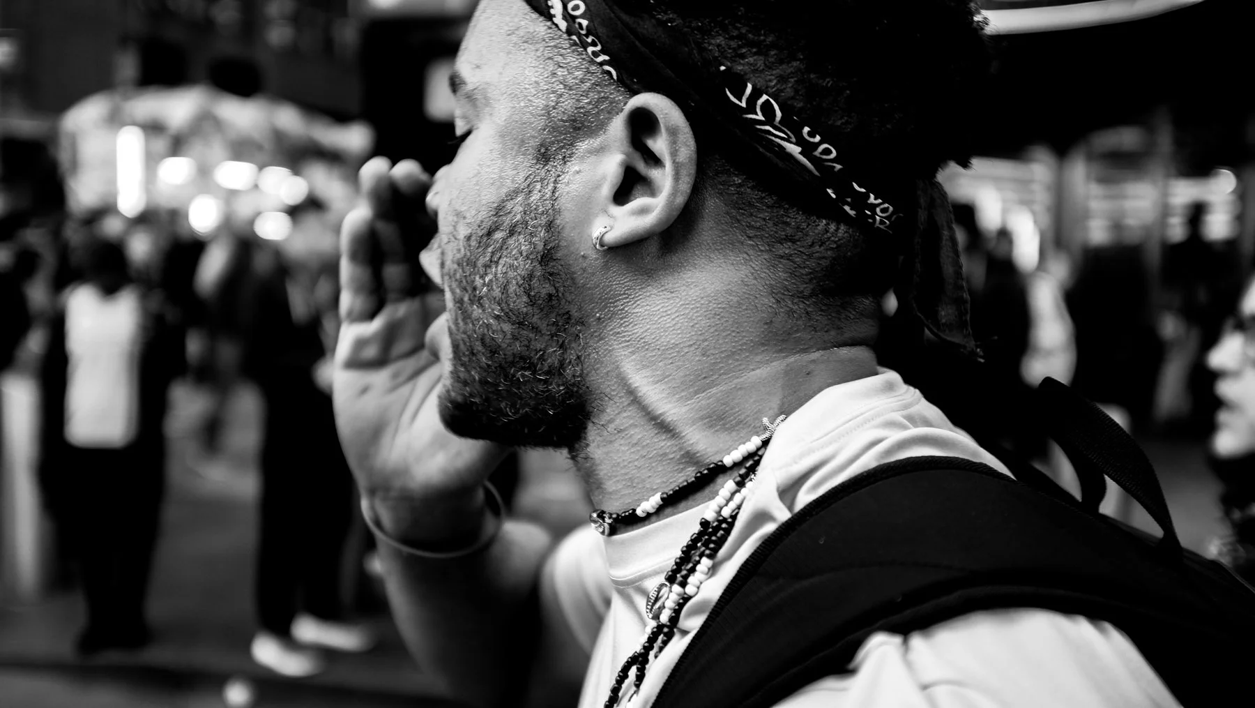 Close-up of a young man's profile with a beard, earrings, and necklace, in a busy outdoor setting with blurred people in the background.