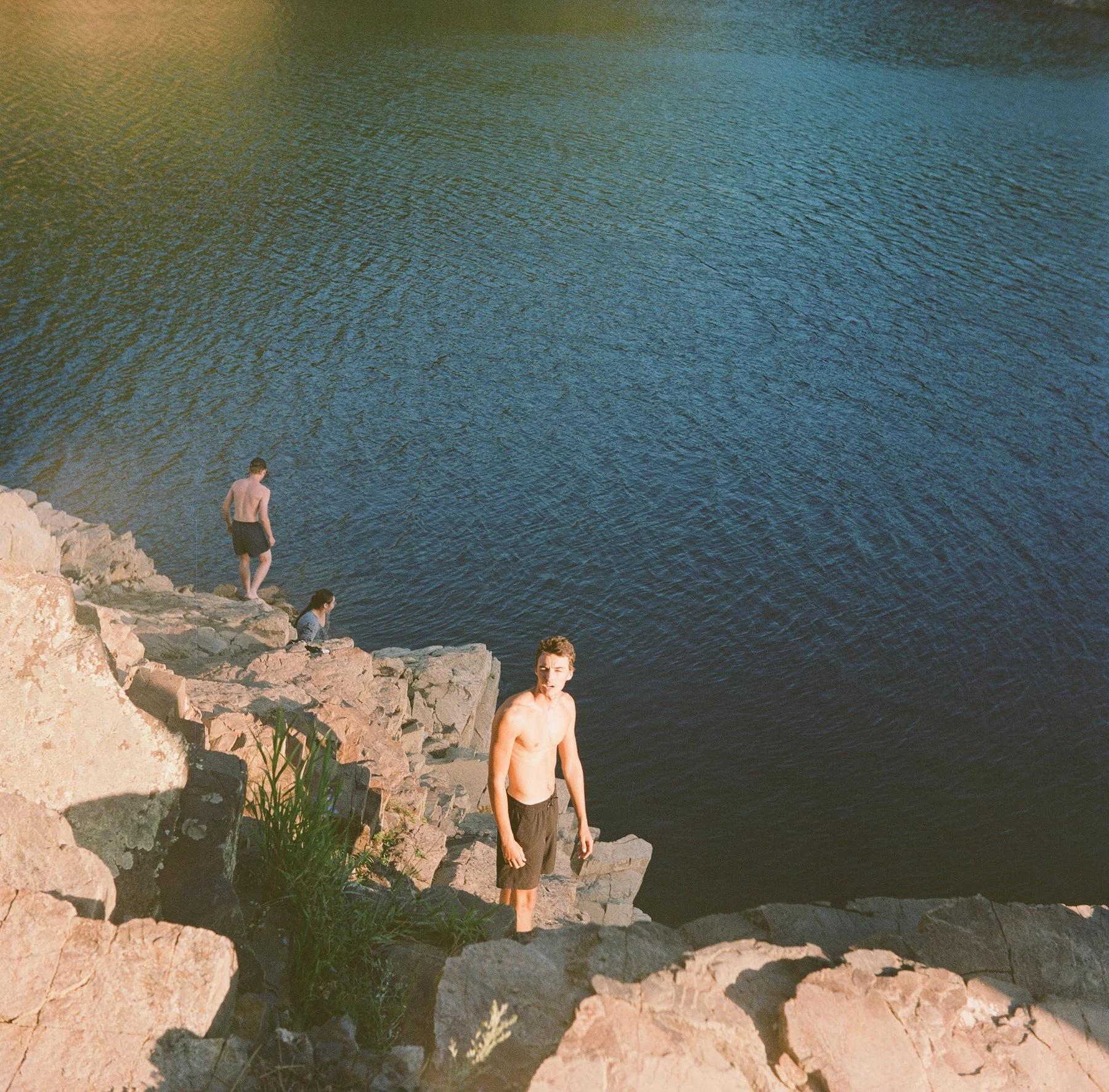 Three young men near a rocky lakeshore with one standing in the water and two sitting or standing on the rocks. One of them is looking at the camera, while the other two are engaged in fishing or relaxing.